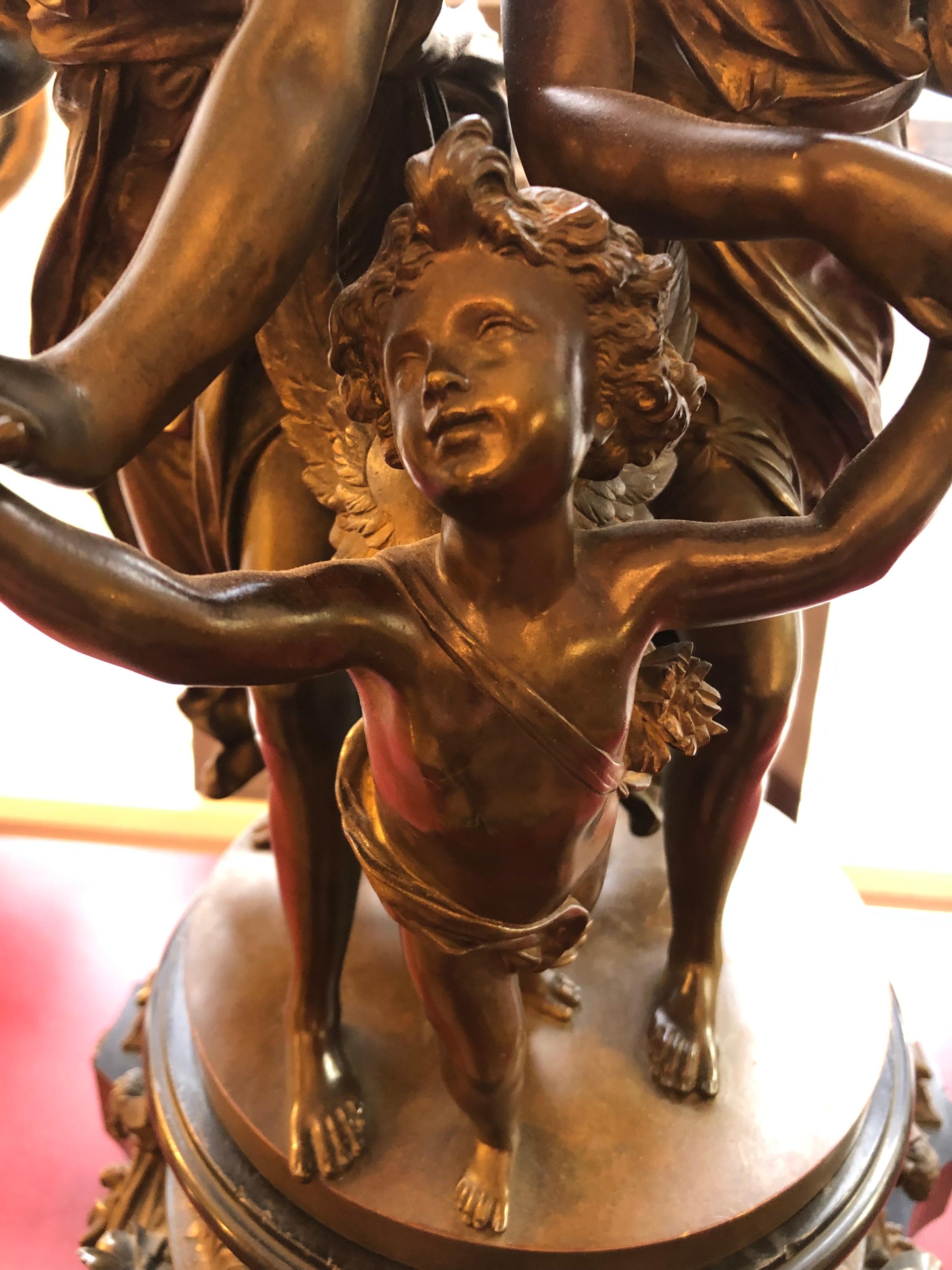 Black marble mantel clock with Dore bronze highlights surmounted by a patinated bronze statue of two partially clad female figures, one holding a tambourine. Both females hold the hand of a cherub, possibly Cupid. All stand on a circular gilt metal