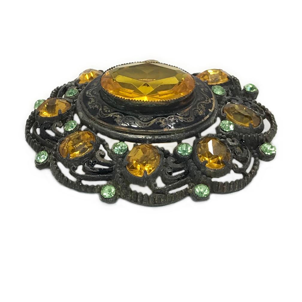 This is a 1930s or 40s scallop edged gunmetal brooch. There is a large bezel set oval shaped amber glass stone surrounded with eight other smaller pieces of prong set oval shaped amber rhinestones. Sixteen smaller peridot colored rhinestones are
