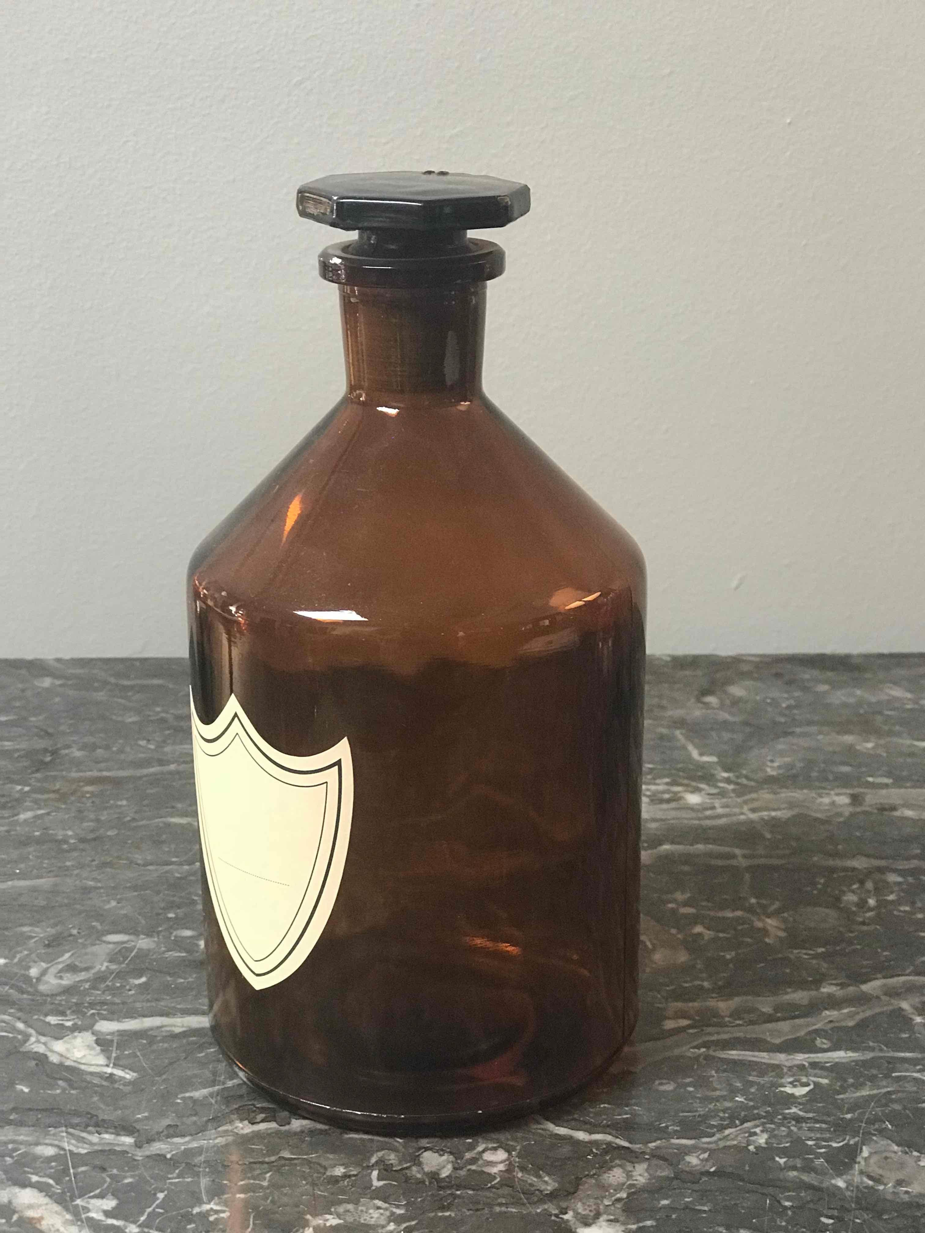 Amber glass apothecary bottle from England circa 1900. Each bottle has a blank cream colored label in the shape of a shield or crest, and is circumscribed with a thin blue border. These are functional objects that could serve as beautiful decorative