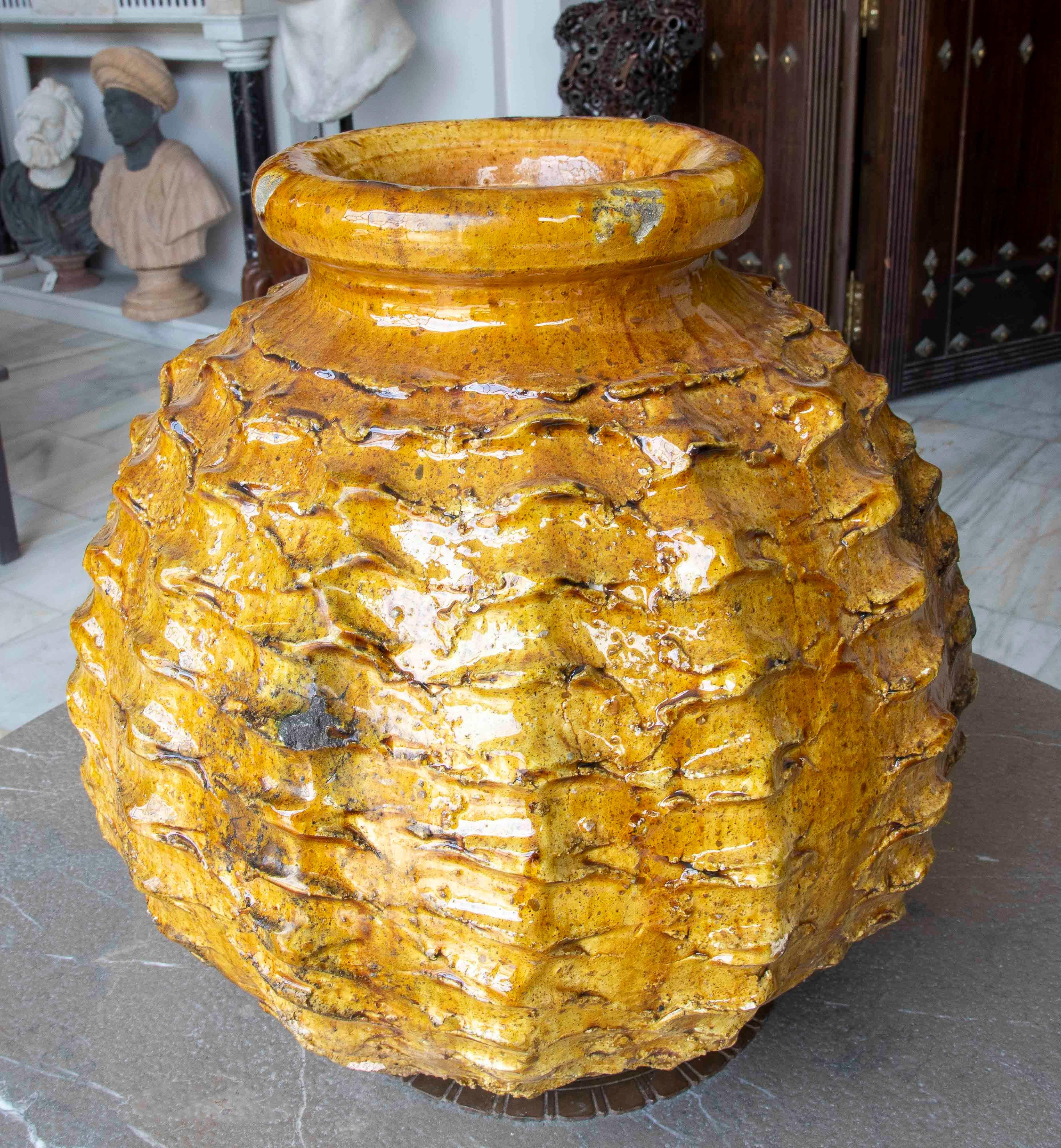 Large artistic ceramic vase, finished in a glossy amber glaze with gold and ochre tones. Its surface features a relief texture reminiscent of natural forms such as bark or exotic fruits. The translucent glaze highlights the irregularities of the