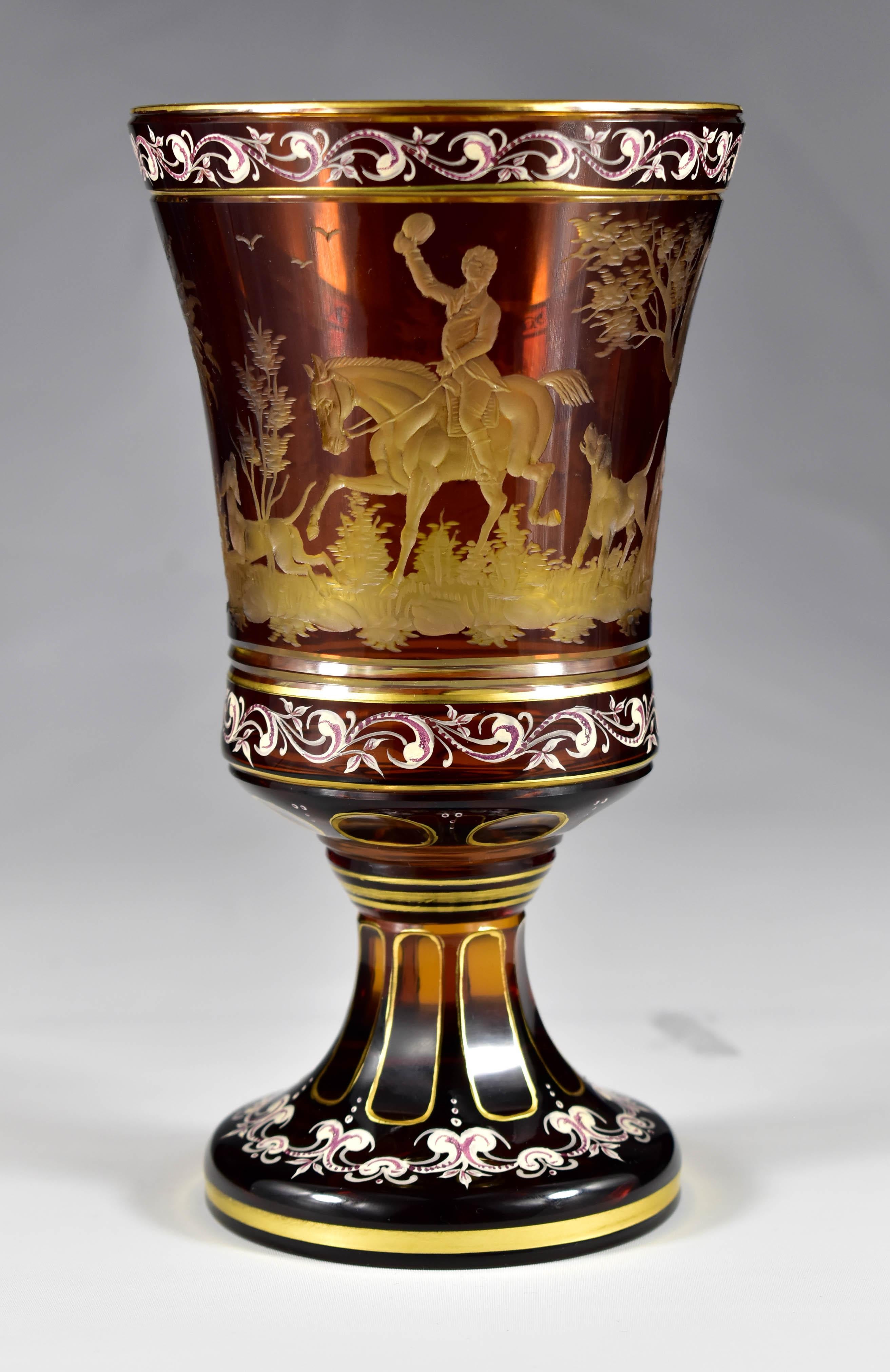 A beautiful goblet made of amber glass overlay with a yellow glaze, decorated with a painting and an engraving of a hunting motif. In the foreground is a rider on horseback with a dog, and in the background are dogs hunting ducks. Everything is