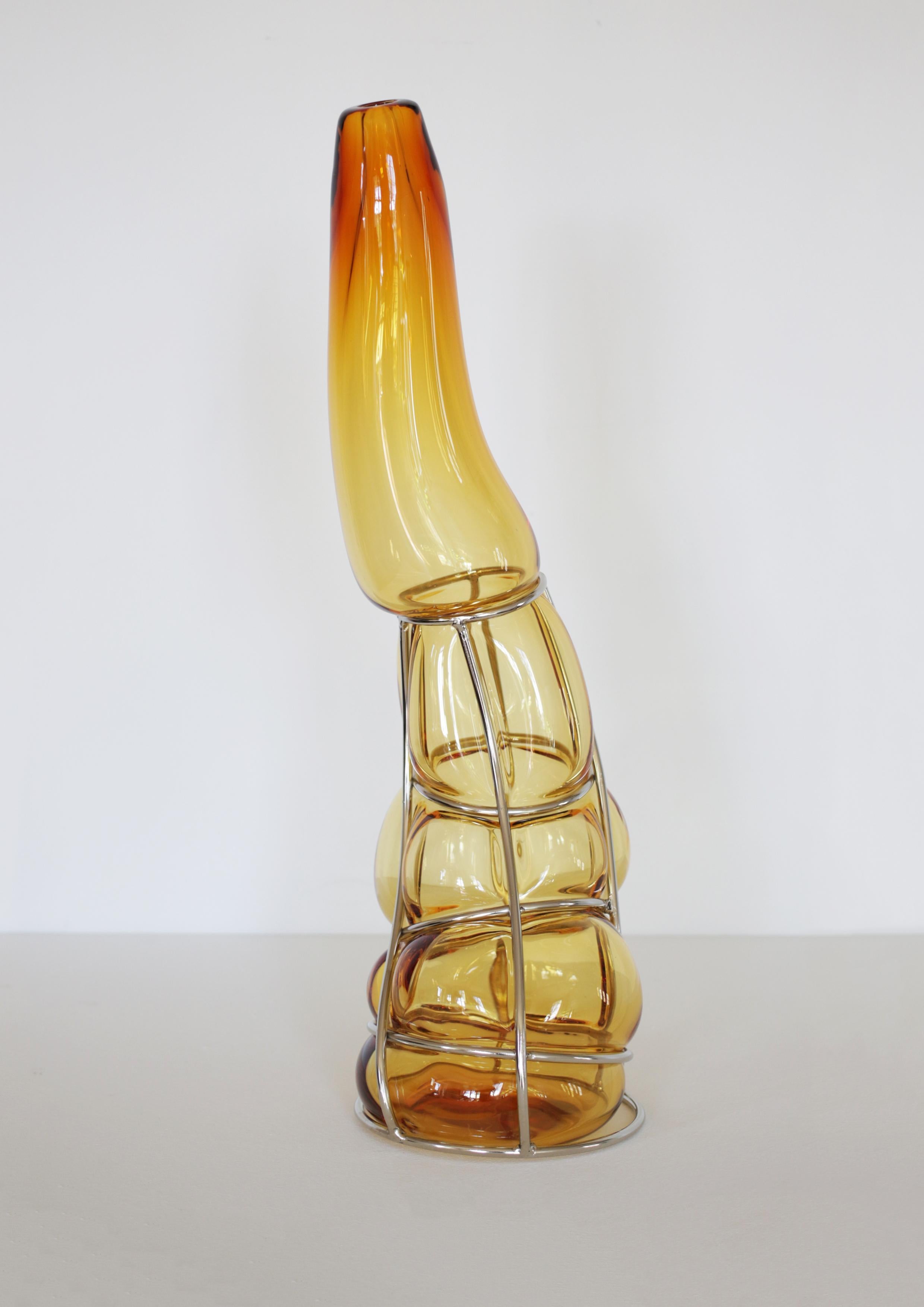 Vaso scultura Ambra Babà di Alice Crepaldi
Un pezzo unico.
Dimensioni: D 20 x L 20 x H 62 cm.
Materiali: Vetro di Murano e metallo.

Questo vaso ingabbiato è caratterizzato da un gambo allungato color ambra con diverse intensità di colore dovute