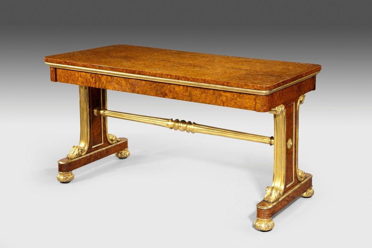 Mesa de biblioteca de la época de la Regencia, de madera de amboyna y dorada, atribuida a Seddon & Seddon. El tablero rectangular está colocado sobre soportes macizos, con un bastidor torneado, dorado y dorado, y patas foliadas doradas, decoradas