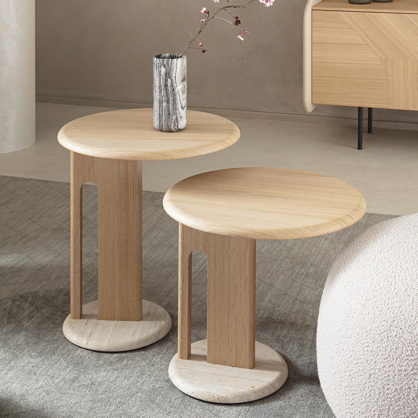 The Ambrogio Basso side table features a medium-sized circular top with a natural oak veneer finished through a membrane press technique. Its distinctive turned oak structure includes an open-arch detail, supported by a solid Roman travertine marble