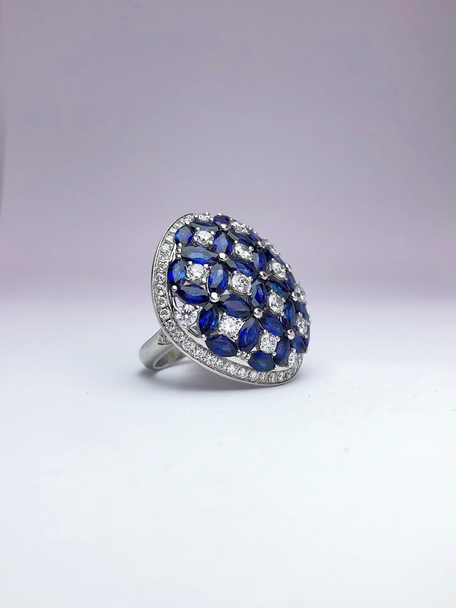 This 18 karat white gold ring is a large cushion shape. The center is uniquely set with blue sapphire marquise cut stones and round brilliant round diamonds. Together the sapphires and diamonds form flower like shapes. The border of the cushion