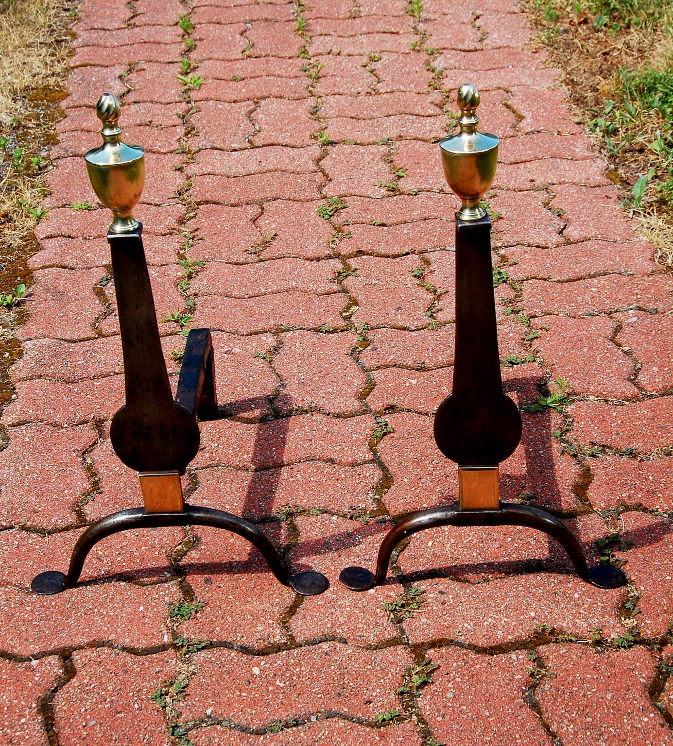American mid 18th century pair of iron knife blade andirons with brass urn and flame finials.  These early andirons are hand wrought iron, embellished with seamed brass urn and flame finials and having a brass rectangular ornamentation just above