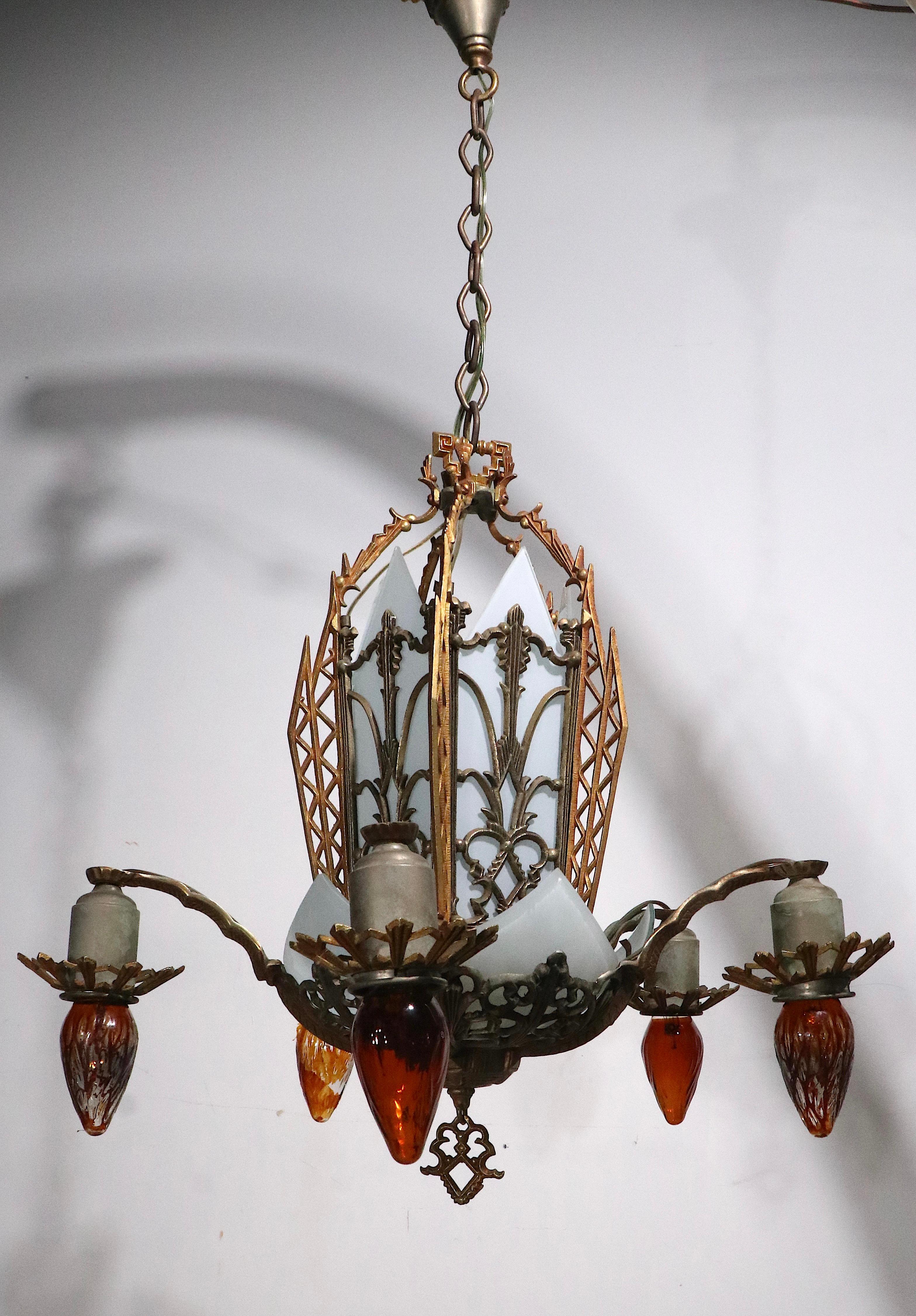 Classic American Art Deco chandelier, having an open work brass frame with original white glass panel shades, curved elements on the bottom, and pointed panels at the top. The fixture has five arms which have down facing exposed bulbs, and a center