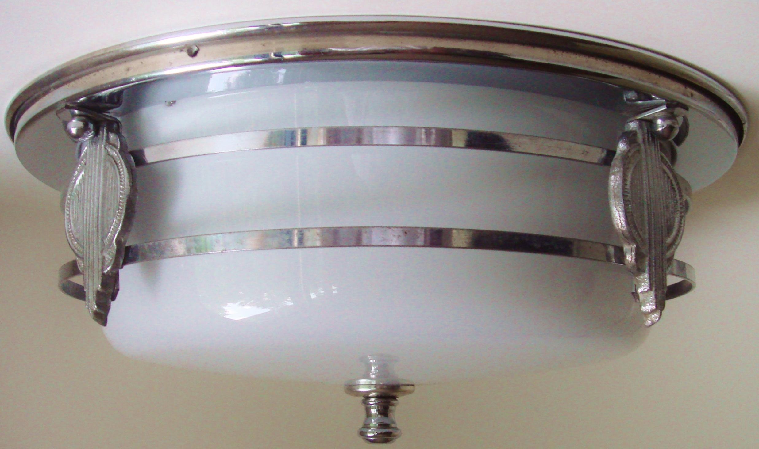 This American Art Deco flush mount ceiling lamp features a milk glass drum shade that is surrounded by a chrome cage. This cage consists of two flat bands of chromed metal that are held together by four ultra Art Deco designed medallions that are in