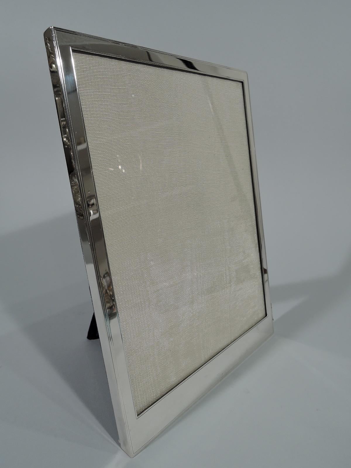 Art Deco sterling silver picture frame. Rectangular window and flat narrow surround with incised wraparound linear borders. Room for engraving at bottom. With glass, silk lining, and velvet back and hinged support. For portrait (vertical) display.