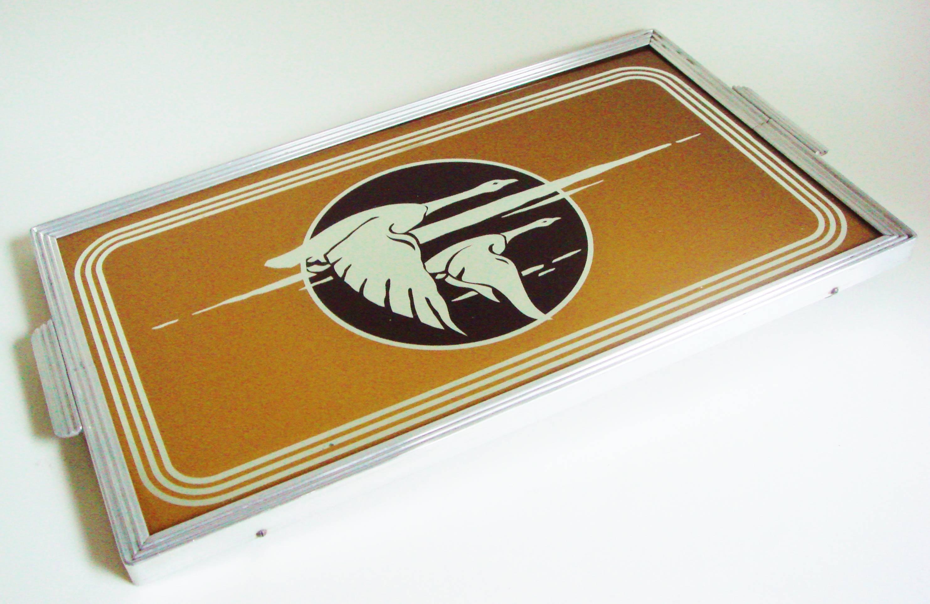 This American Art Deco reverse painted glass figural cocktail tray features two mirrored geese flying over a black sun with mirrored clouds and framed by three mirrored bands. The sand colored background is surrounded by a ribbed aluminum frame with