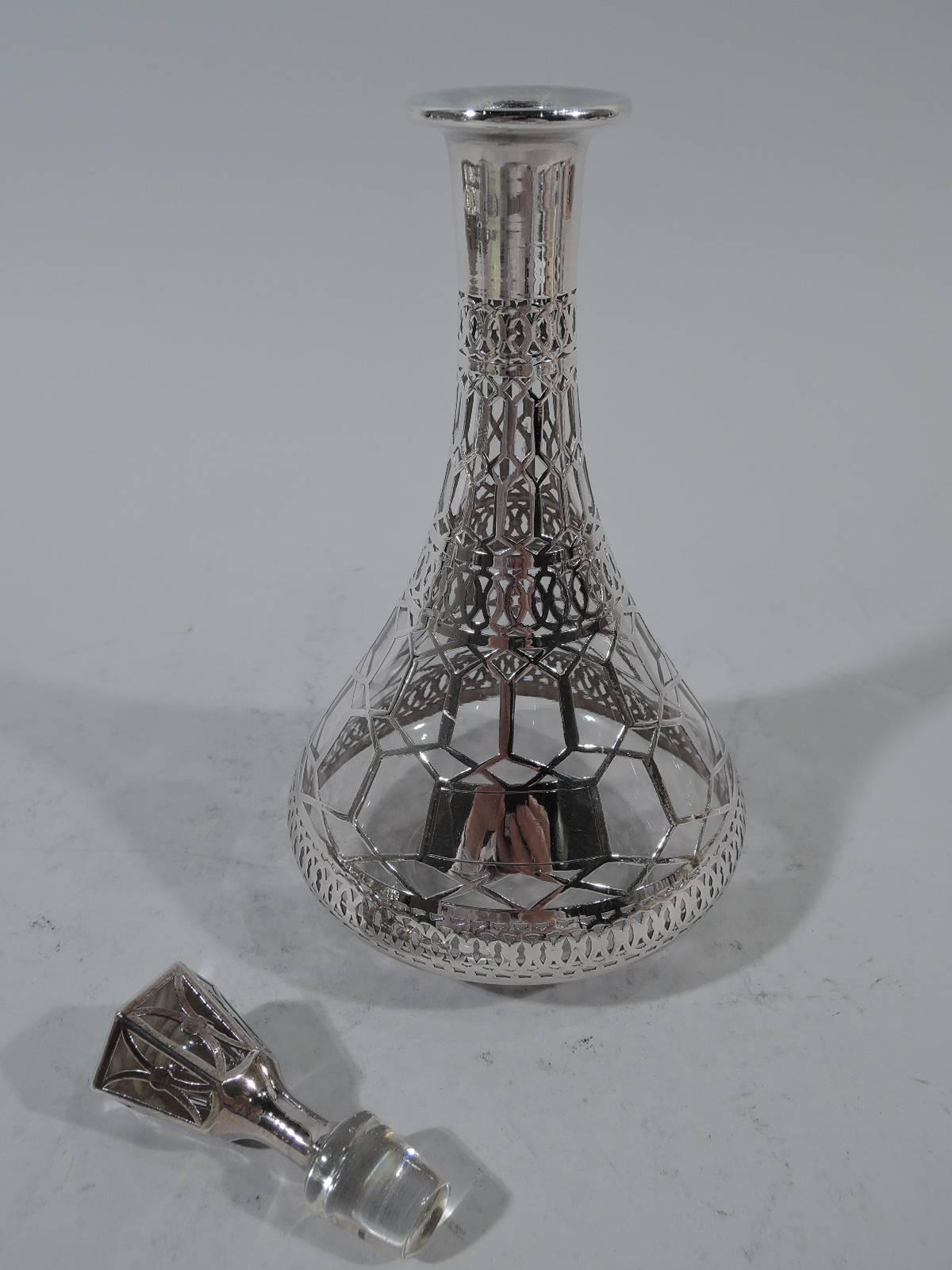 Art Deco clear glass decanter with silver overlay. Conical bowl, cylindrical neck, and flat everted rim. Stopper tapering and hexagonal with short plug. Interlaced geometric ornament with circles and hexagons. Vacant rectangular frame. Also vacant
