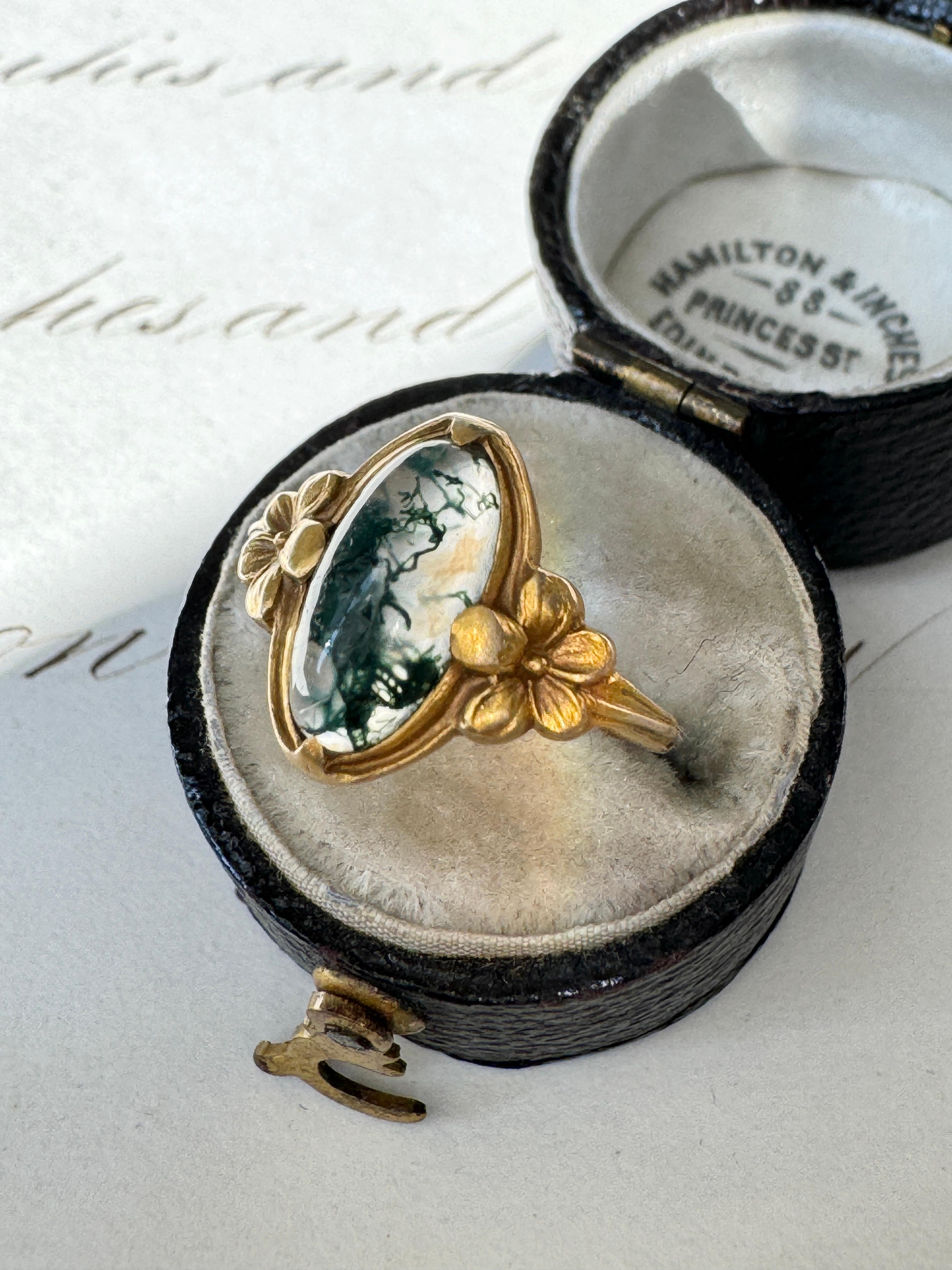 This sweet little Art Nouveau pinky ring by Jones & Woodland Co. of Newark, NJ features an elongated moss agate cabochon presented in a 14 karat gold mounting. The shoulders are artfully adorned with a pair of delicate flowers. Currently a ring size