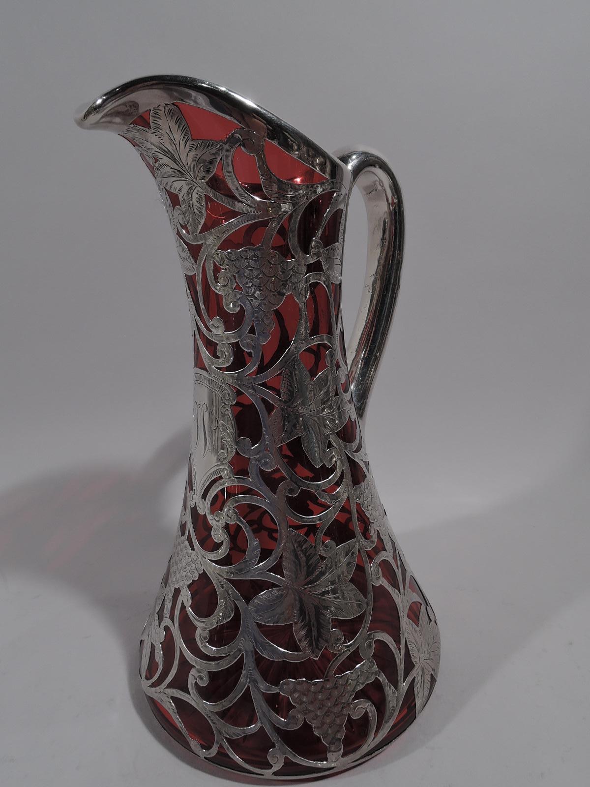 Turn-of-the-century American Art Nouveau red glass pitcher with engraved silver overlay. Conical with helmet mouth and looping scroll handle. Fruiting grapevine overlay with loose, rambling scrolls, oversized leaves, and irregular, semi-abstract