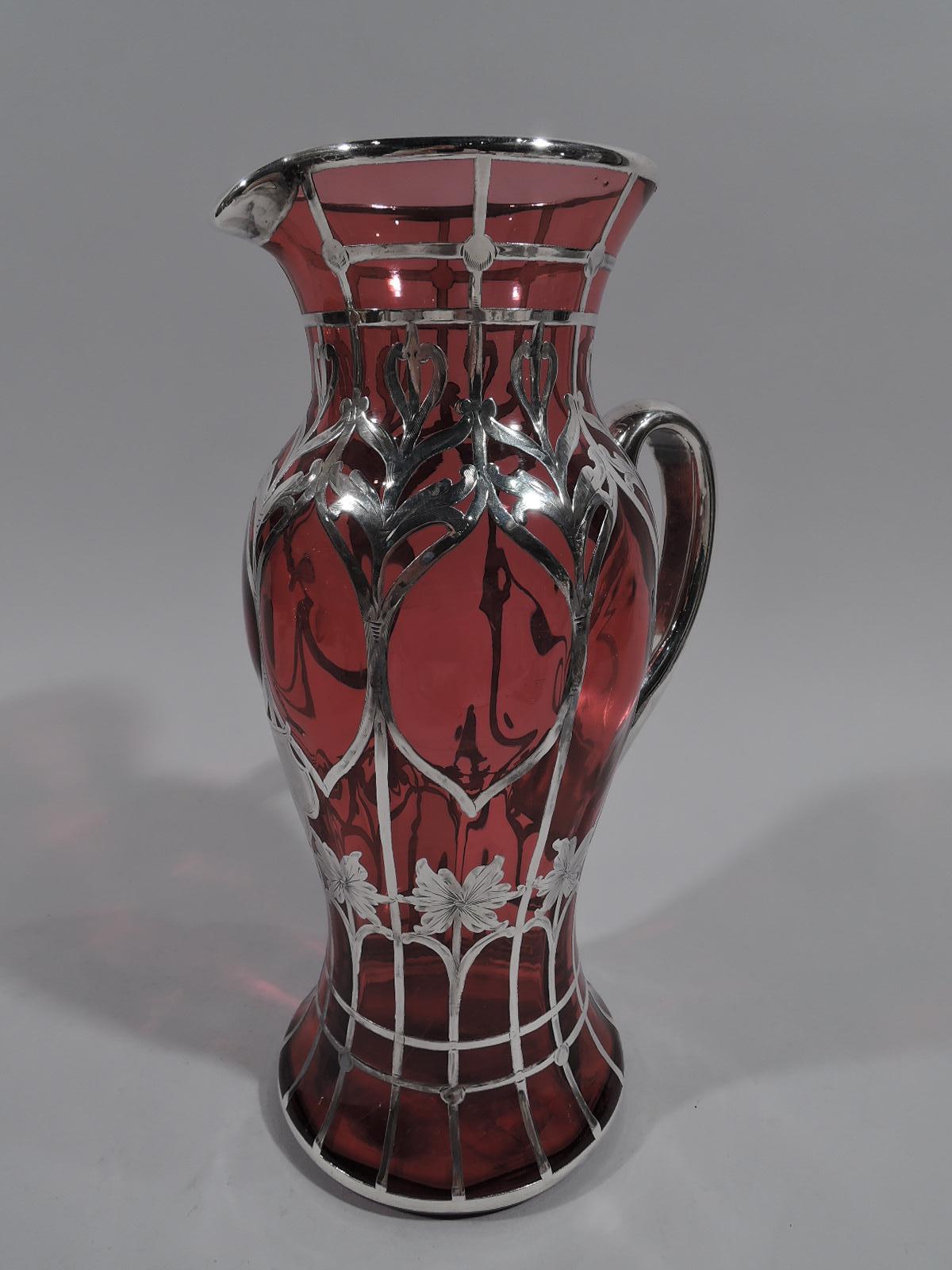 Turn of the century American Art Nouveau red glass claret jug with engraved silver overlay. Baluster with spread foot, looping handle, and lip spout. Vertical trellis-style grid with stylized stem flowers and leaves. A controlled rectilinear pattern