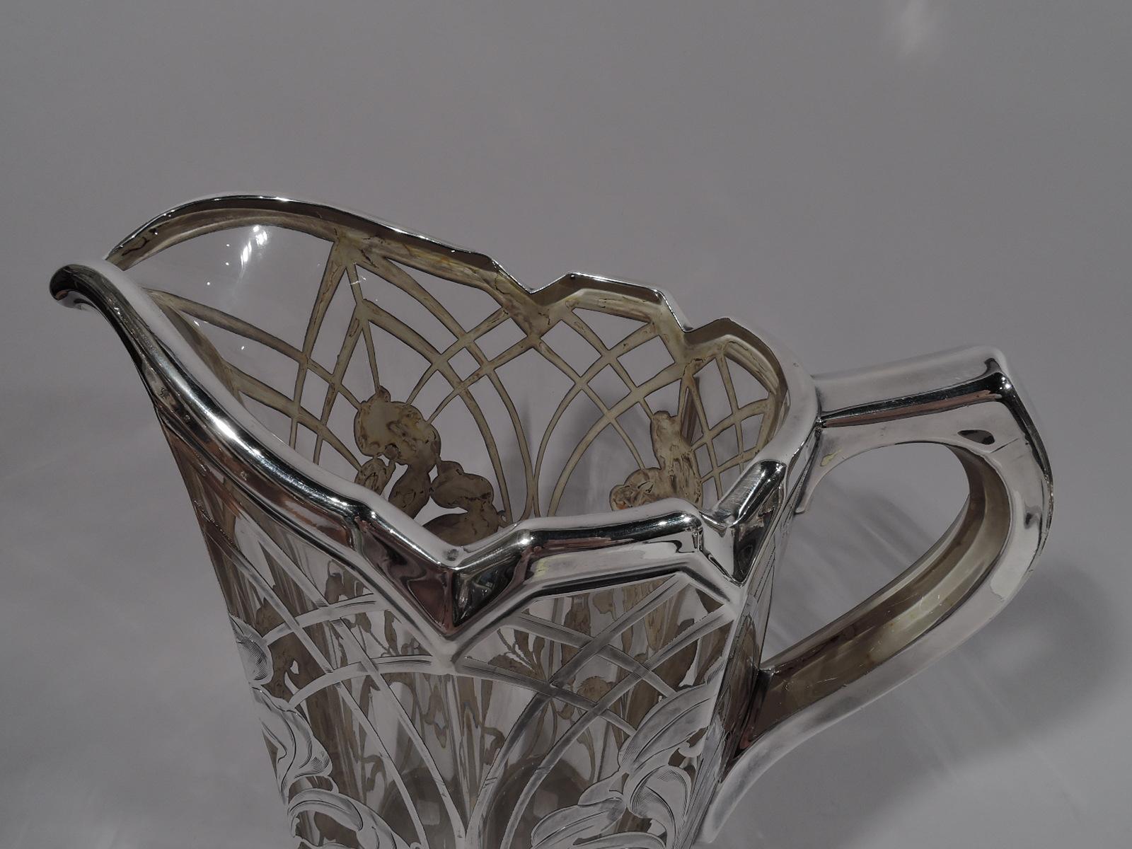 Large turn-of-the-century American Art Nouveau water pitcher. Faceted clear glass body with asymmetrical and castellated helmet mouth. Silver overlay in form of trellis arcade inset with loose and fluid irises. Scroll handle in silver collar as is