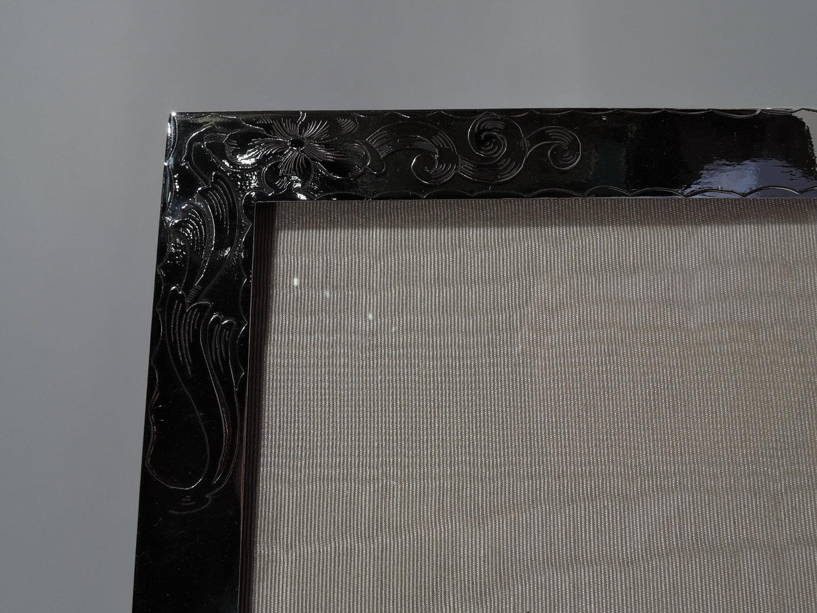 Turn-of-the-century American Art Nouveau sterling silver picture frame. Rectangular window in flat surround. Loose and fluid acid-etched scrolls and flowers between wave borders. With glass, silk lining, and velvet back and hinged support for