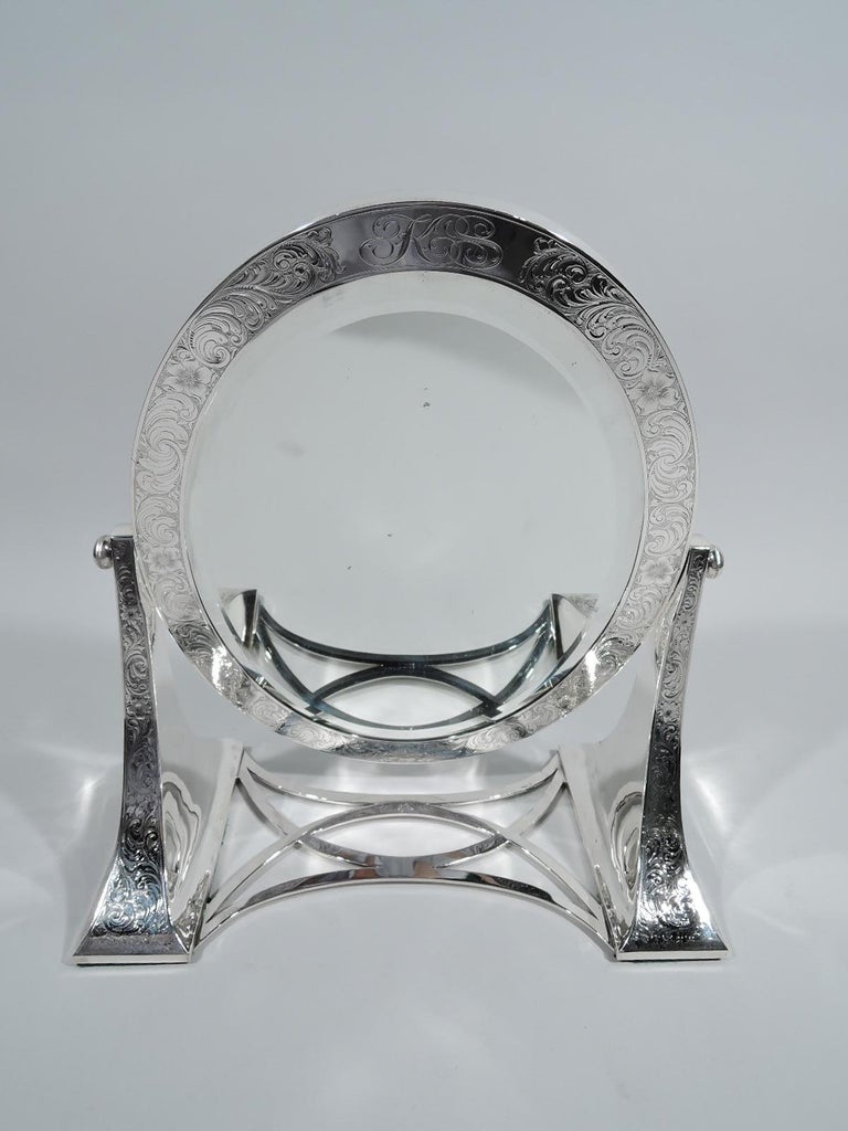 Turn-of-the-century Art Nouveau sterling silver mirror. Made by Lebkuecher in Newark. Two bracket supports joined by concave and crisscrossing stretchers. Hinged swing oval frame inset with bevelled glass. Dense and fluid engraved scrolls, leaves,