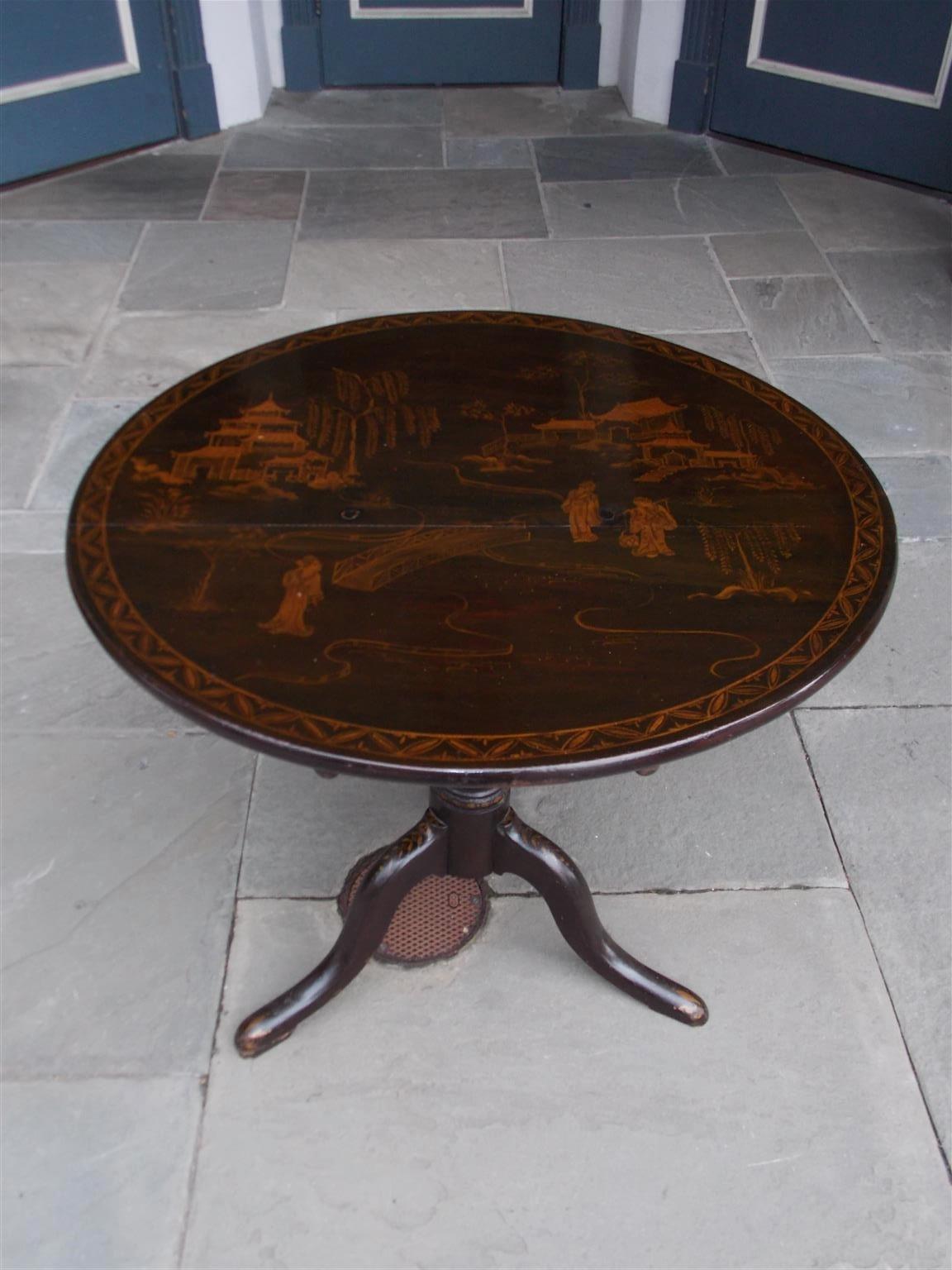 American black lacquer Japanned tilt top table depicting a landscape scene with pagodas in the background, a flowing stream with figures in the foreground, retaining the original locking mechanism and terminating on a turned ringed tripod pedestal