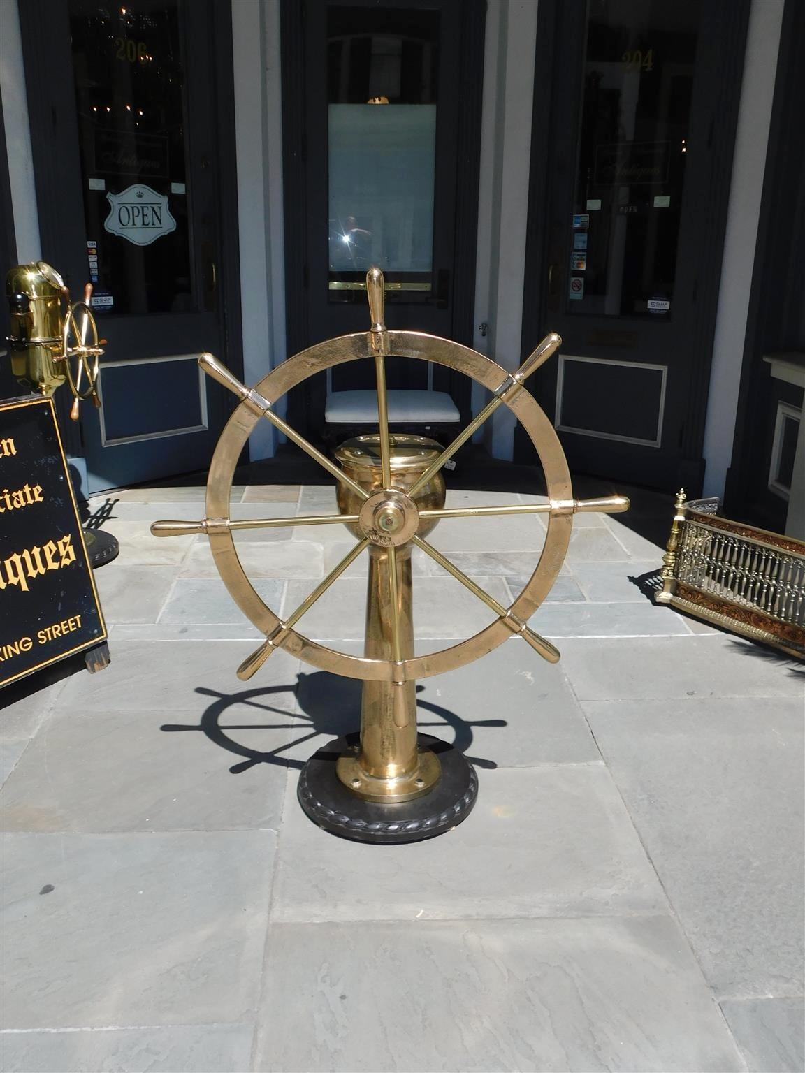 American brass nautical ship wheel with a central circular brass hub, eight brass spindles with bulbous handles, solid brass geared pedestal, rudder indicator arrow, and mounted on a circular carved rope base. Late 19th century. Stamped Dake Engine