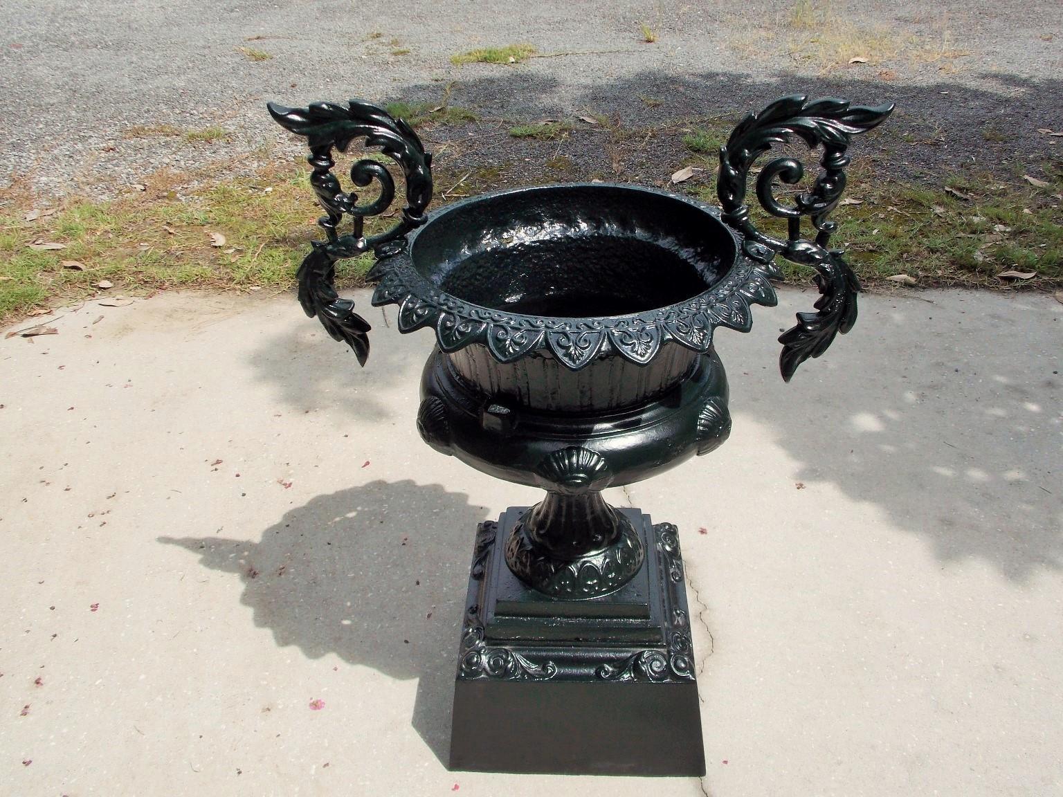 American cast iron and powder-coated campana garden urn with a triangular floral molded edge, flanking acanthus scrolled side handles, circular medallions, and resting on fluted squared floral decorative step back plinth base, Mid-19th century. Urn
