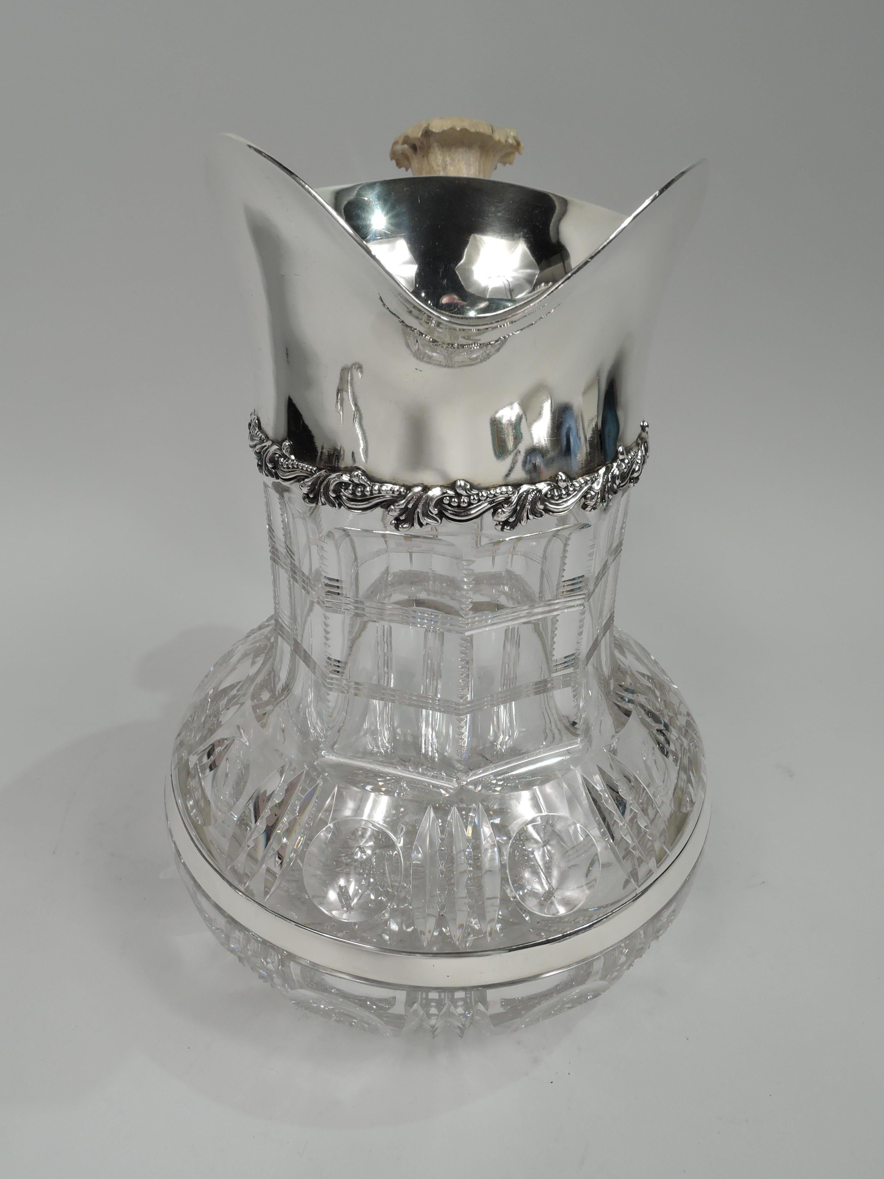 Edwardian Classical water pitcher. Made by Redlich in New York, ca 1890. Clear cut-glass with bead-and-reel bands on bowl and brilliant star on underside; Neck faceted and notched with reeded bands. Sterling silver collar with helmet mouth and