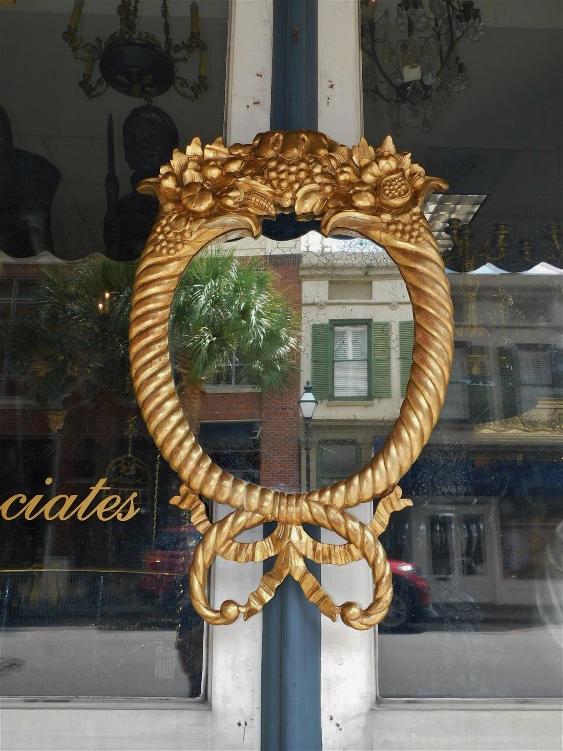 American Classical gilt carved wood Cornucopia and grape cluster oval wall mirror with flanking scrolling terminals bound by a lower intertwined ribbon pendent, Early 19th Century. Mirror retains the original silvered glass and wood backing. 