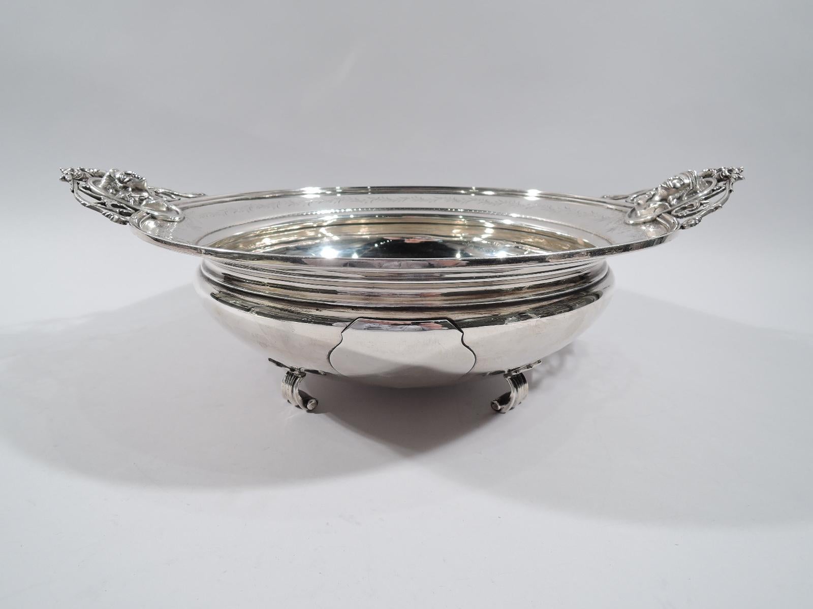 American sterling silver centerpiece bowl, circa 1870. Bellied with applied cartouche (vacant) and 4 shell-mounted scroll supports. Tapering shoulder engraved with scrolling leaf border. Open scrollwork side handles inset with cast medallion heads.