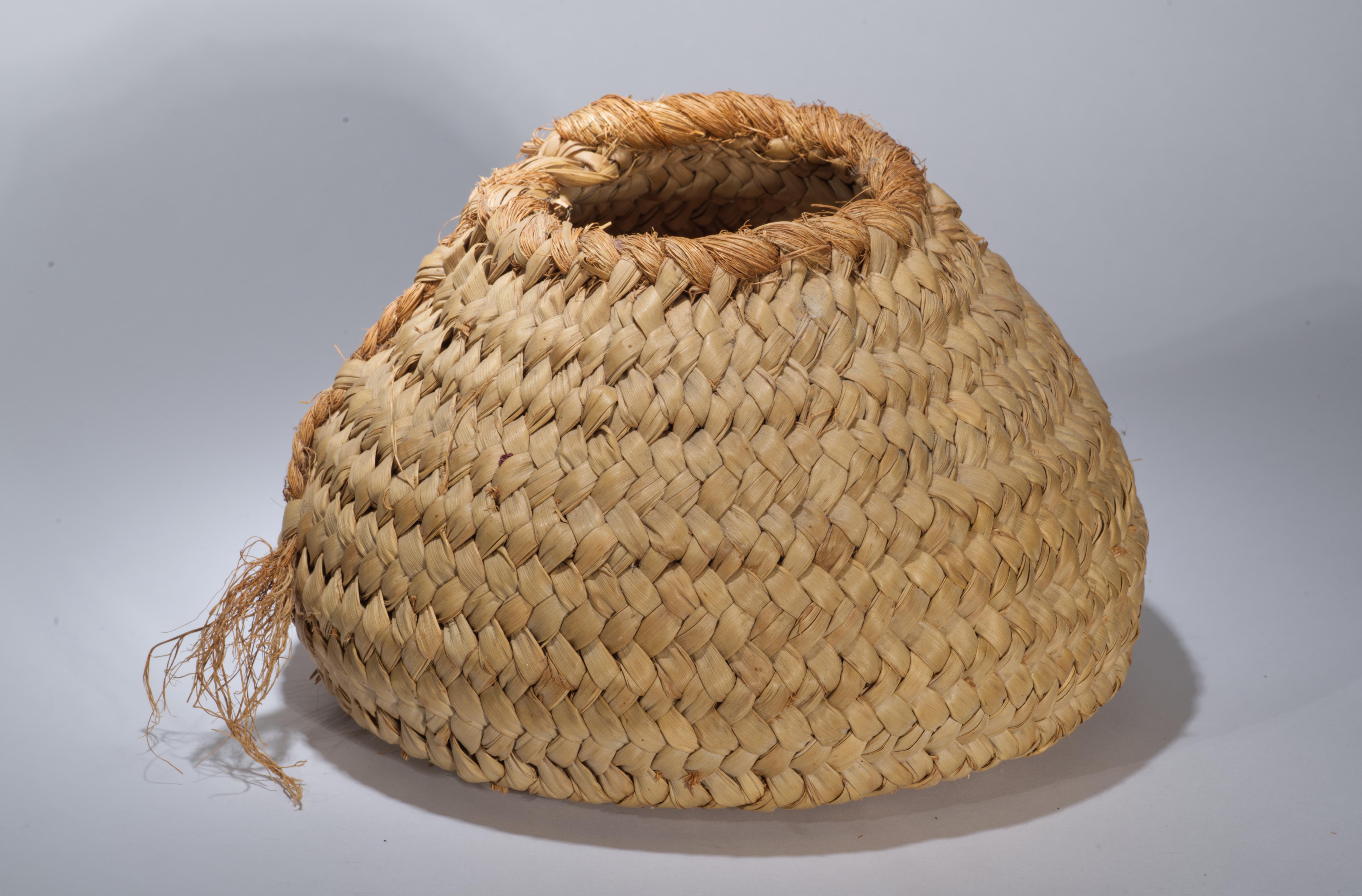 The vintage basket features rounded, bulbous form with wider base and tapered top. The top is accented with decorative woven grass elements that are extrended to the body of the basket, creating a free-flowing, asymmetrical textured decor. The