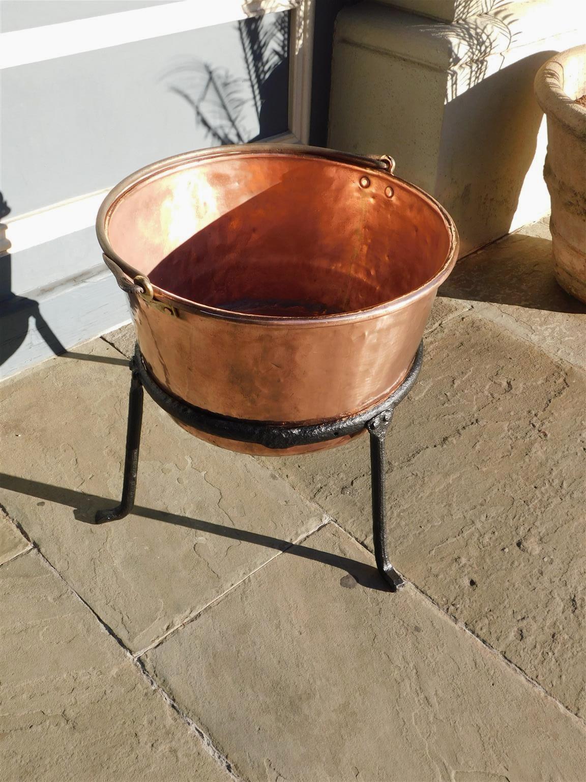 American copper and wrought iron plantation cauldron with brass ring mounts, original wrought iron folding handle, and supported by the original circular fitted three leg Stand with stylized feet. Late 18th century.