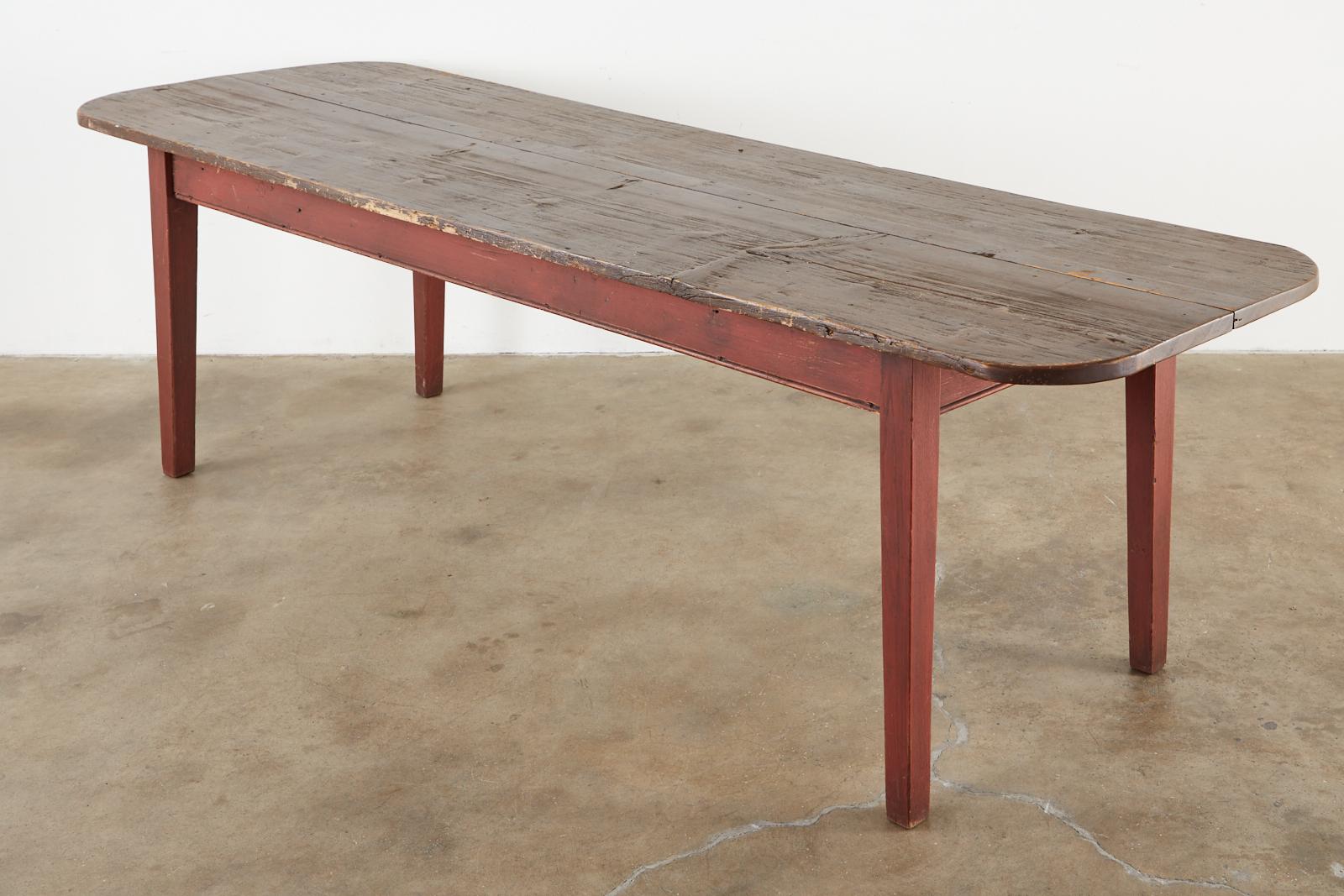 Rustic American country farmhouse harvest table or dining table crafted from reclaimed pine wood. The top is made from two 1 inch thick long planks having rounded ends. The base is painted barn red and is supported by square, tapered legs. Ample leg