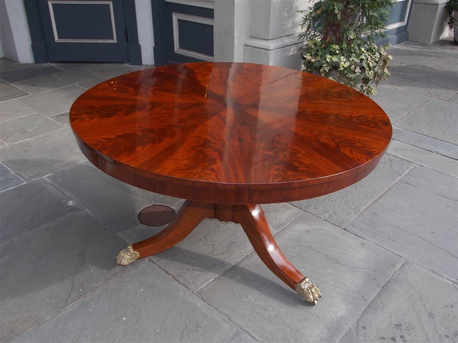 American Honduras crotch mahogany hinged center table with pie shaped top, banded skirt, original brass locking mechanism, turned bulbous pedestal, and resting on three molded splayed legs with the original hairy paw brass feet on casters. New York,