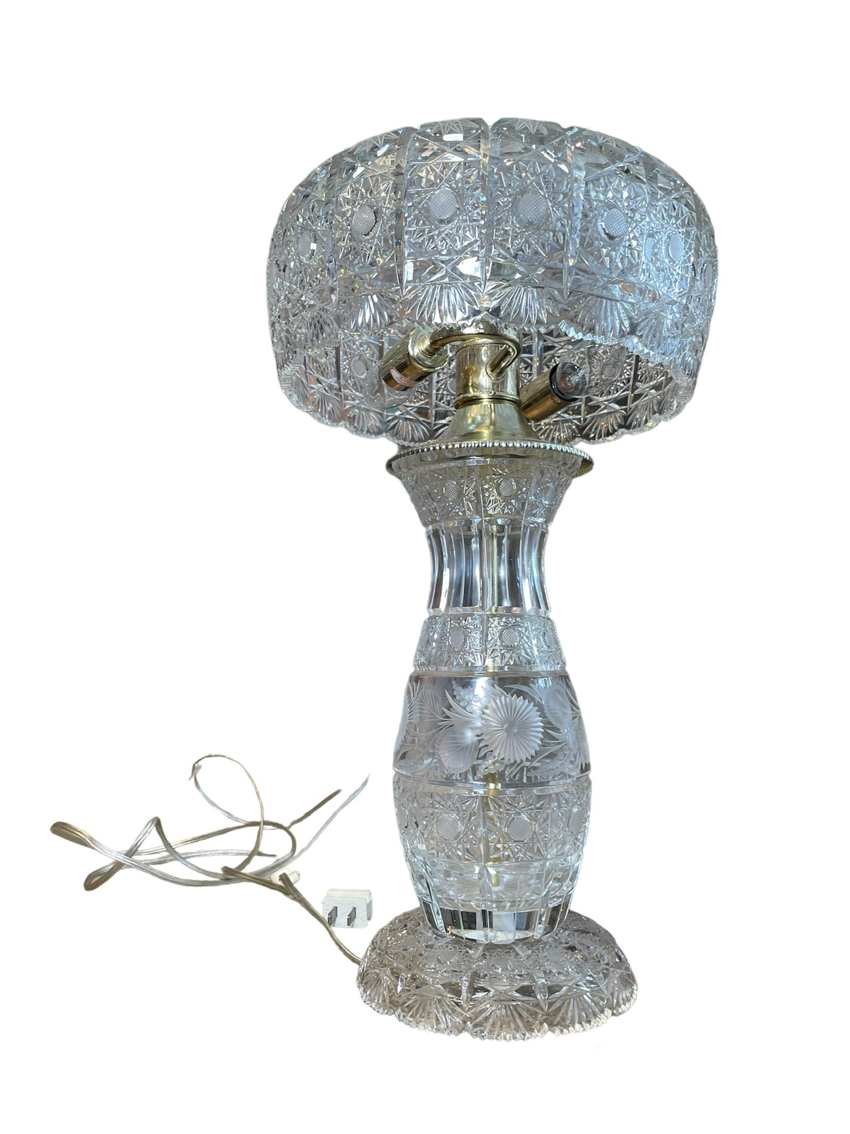 This is an American Cut Crystal Electrified Table Lamp. It depicts a round wide bowl shaped cut crystal shade supported by an also cut crystal urn shaped base/body. Both cut crystals depict a pattern of stars, starbursts and fans. The center of the