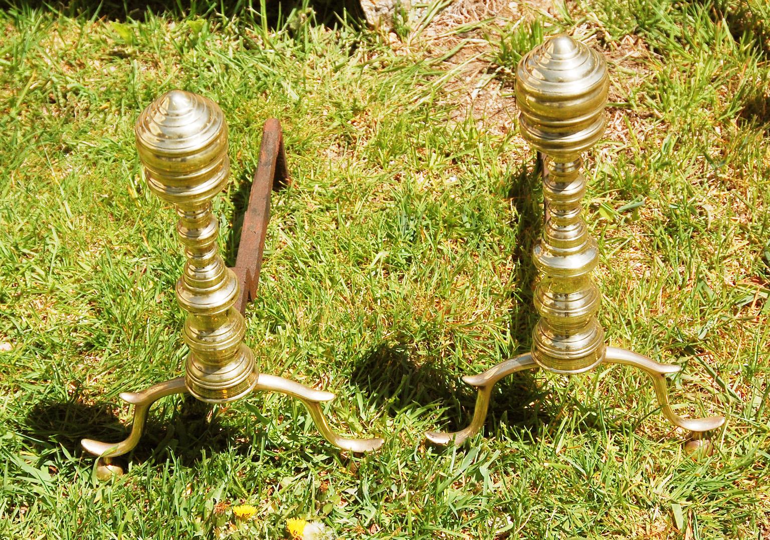 American Empire period brass andirons with cabriole legs, spurred knees, sitting on ball feet. The wrought iron log holders are stepped down so there was no need for brass log stops, the shape of the log holders kept the logs from impinging on the