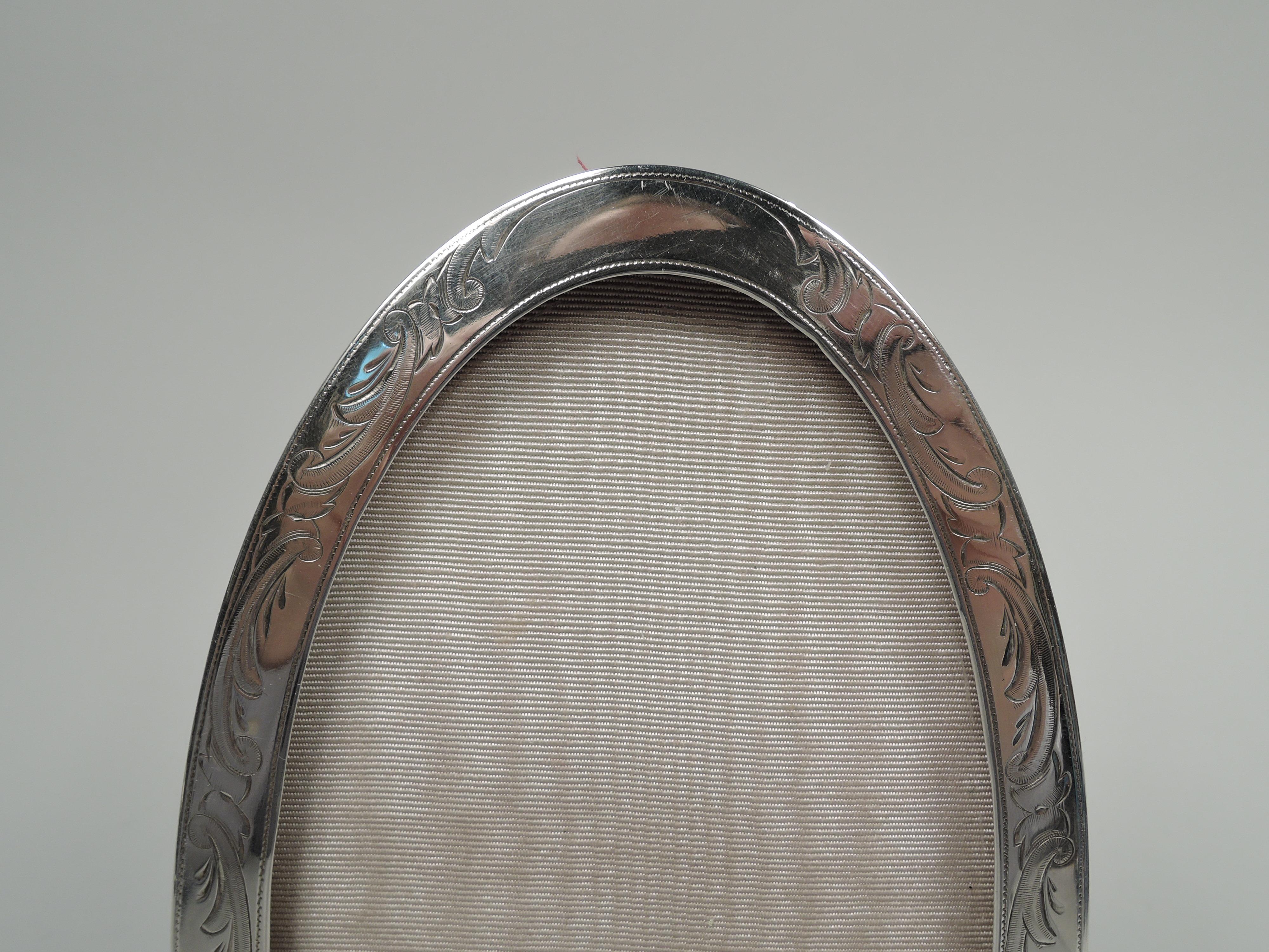 Edwardian Classical sterling silver picture frame. Made by Mauser in New York, circa 1900. Oval window in flat surround on open triangular supports. Engraved leafing scrolls and pendant flowers, and pointillé borders. With glass, silk lining, and