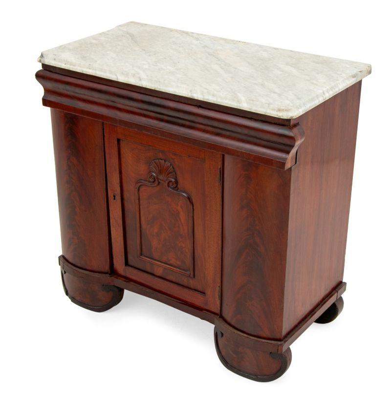 Armadietto American Empire in mogano fiammato con piano in marmo, con un lungo cassetto sopra un'anta singola che si apre su due ripiani, l'anta intagliata con motivo a conchiglia. 29,5