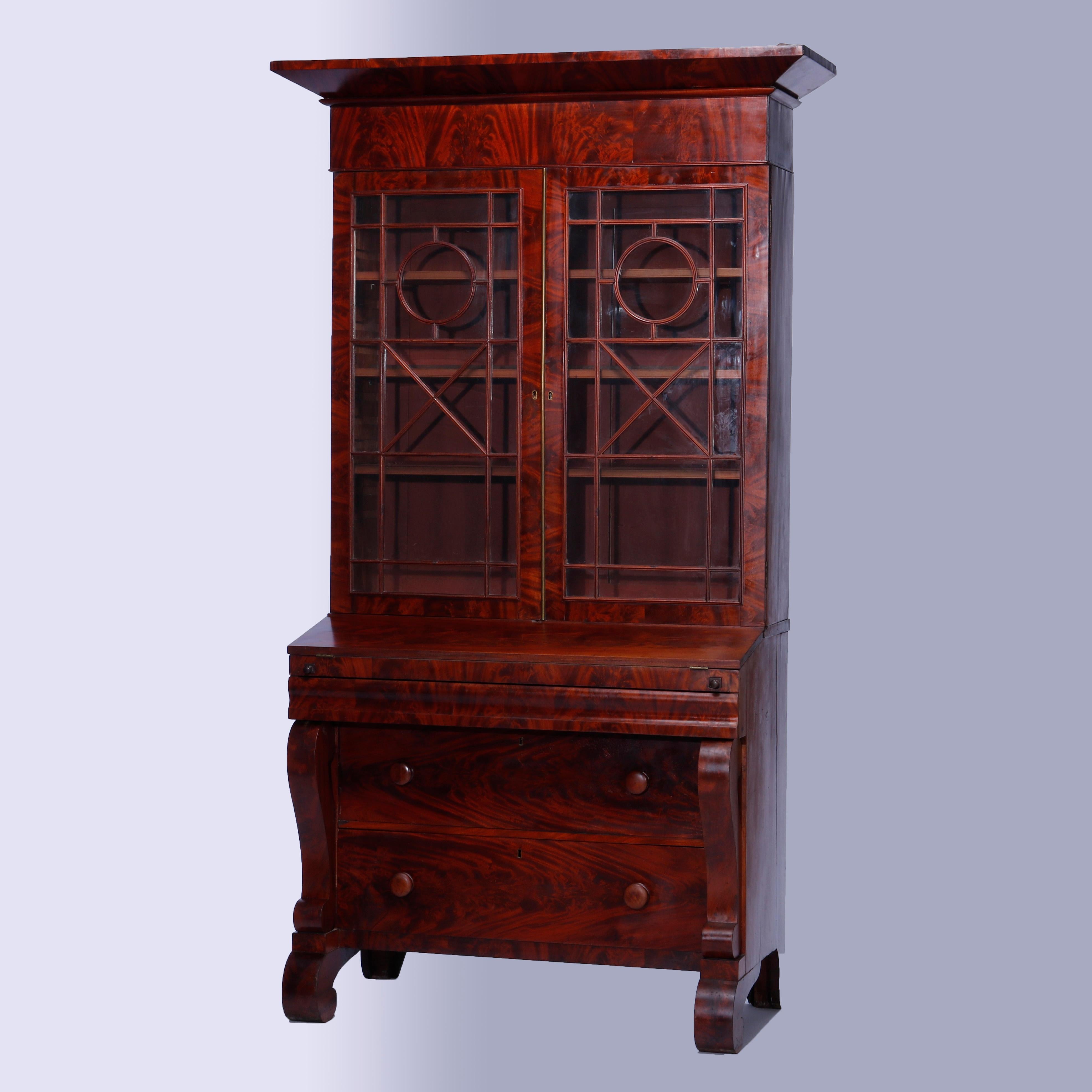 An antique American Empire neoclassical secretary in the manner of Joseph Meeks offers flame mahogany construction with upper bookcase having double glass doors opening to shelved interior and surmounting lower case with drop-front desk and ogee