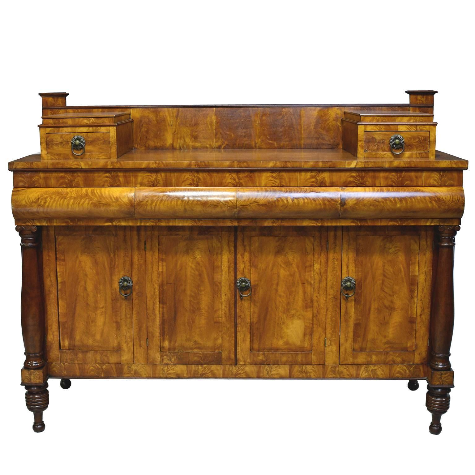 A handsome American Empire sideboard in fine satinwood with two small top drawers and a backsplash over a base with three convex drawers above four cabinet doors that are flanked at each end by a turned column. Base rests on ring-turned feet.