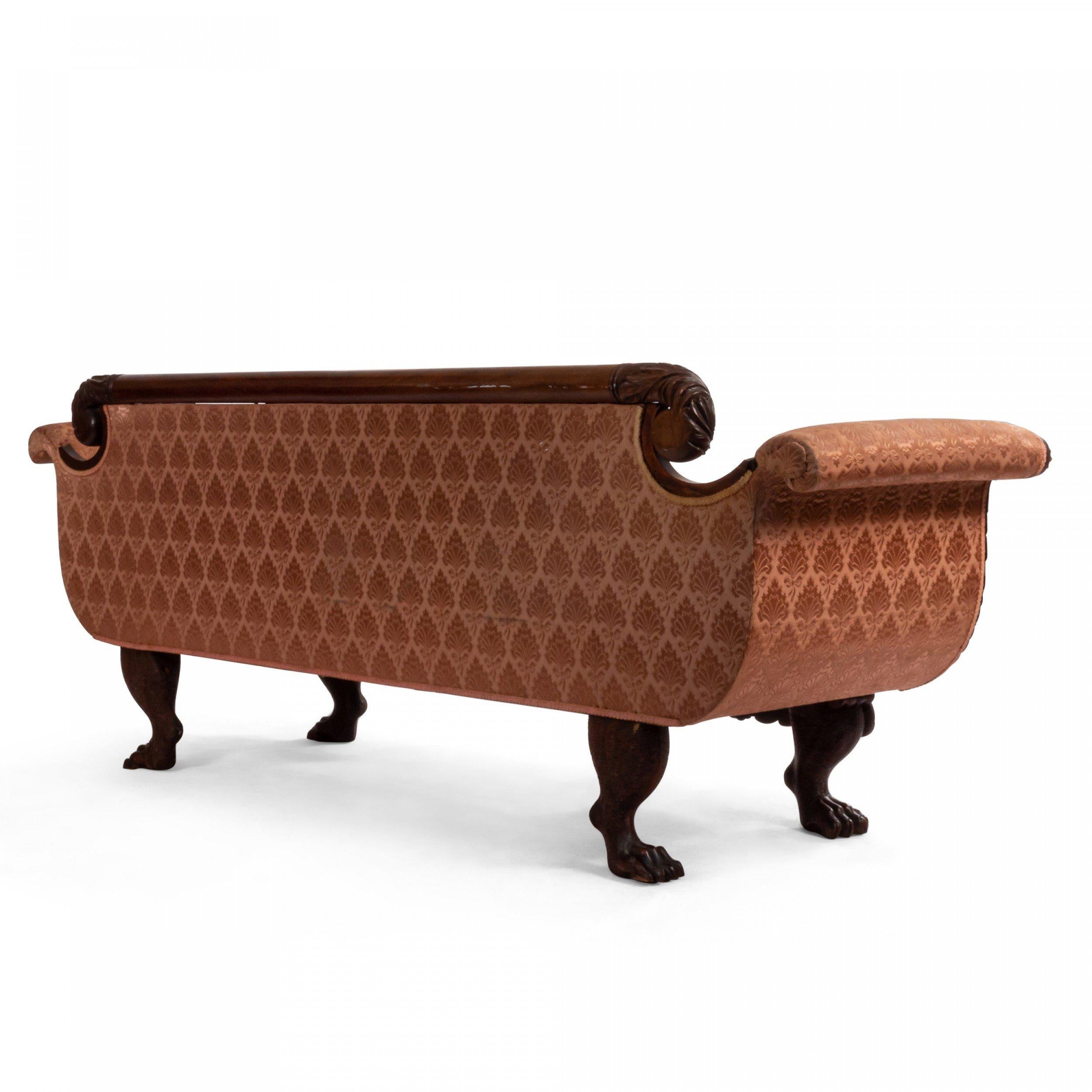 American Empire style (19th century) peach upholstered sofa with matching bolster pillows and a mahogany frame with scrolled arms and lions paw feet with a relief carving of cornucopia decorations at legs. 
  