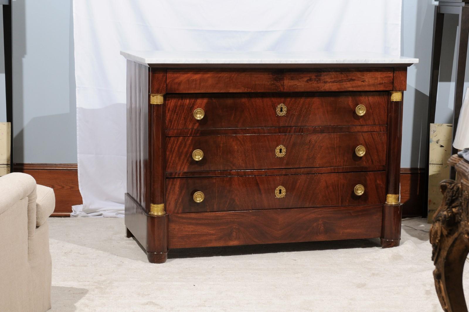 Early 20th century mahogany Empire style chest of drawers or dresser, having an overhanging white marble top and cornice, supported by flanking applied columns on a rounded bases, with an undecorated apron, bun feet, and gilt metal escutcheons,