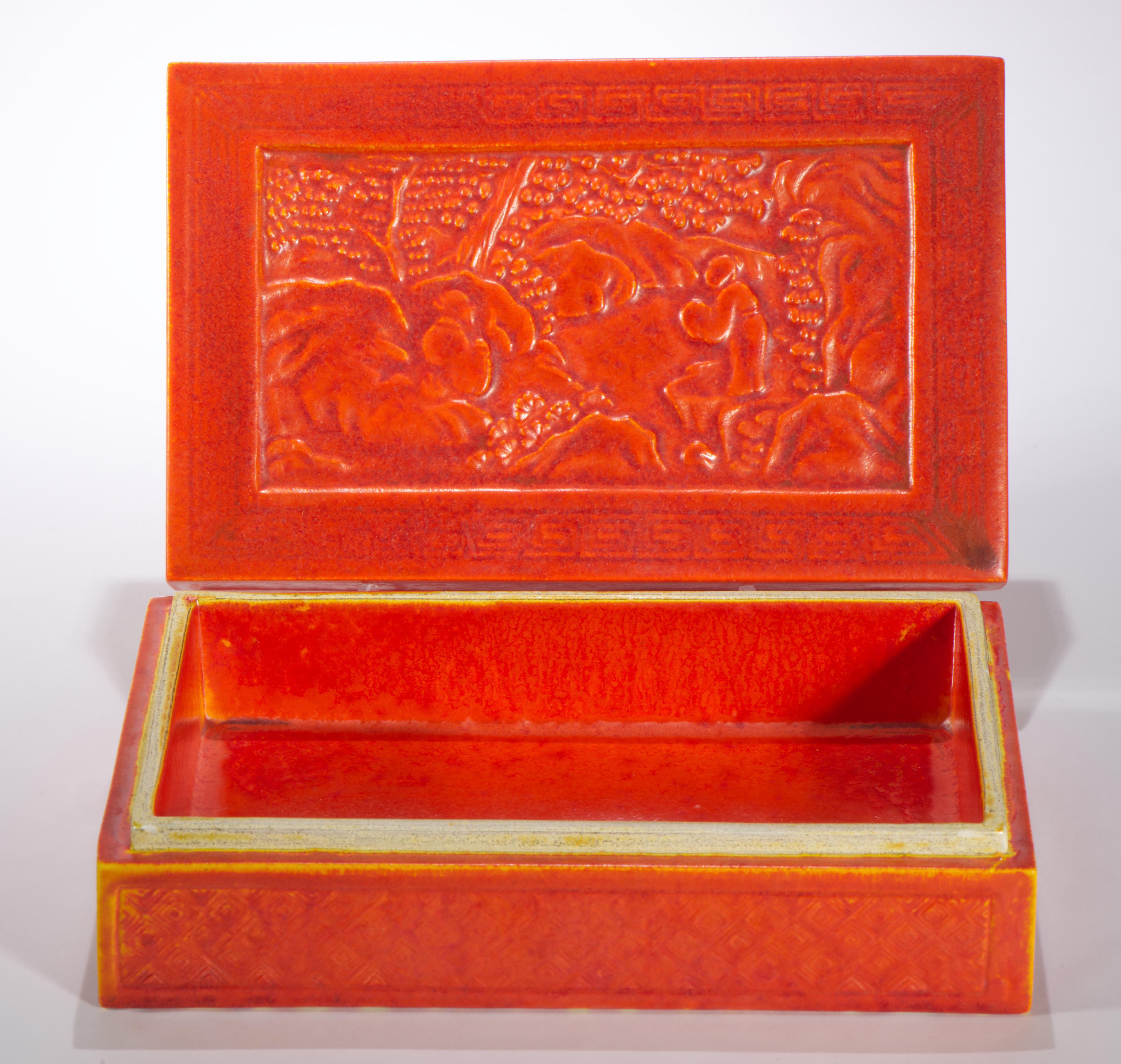 The unusual decorative ceramic box features complex Chinoiserie style decor in relief, with bright orange, cinnabar colored semi-matte glaze. The central design on the lid depicts human figures in Asian clothes, standing in a garden or rural