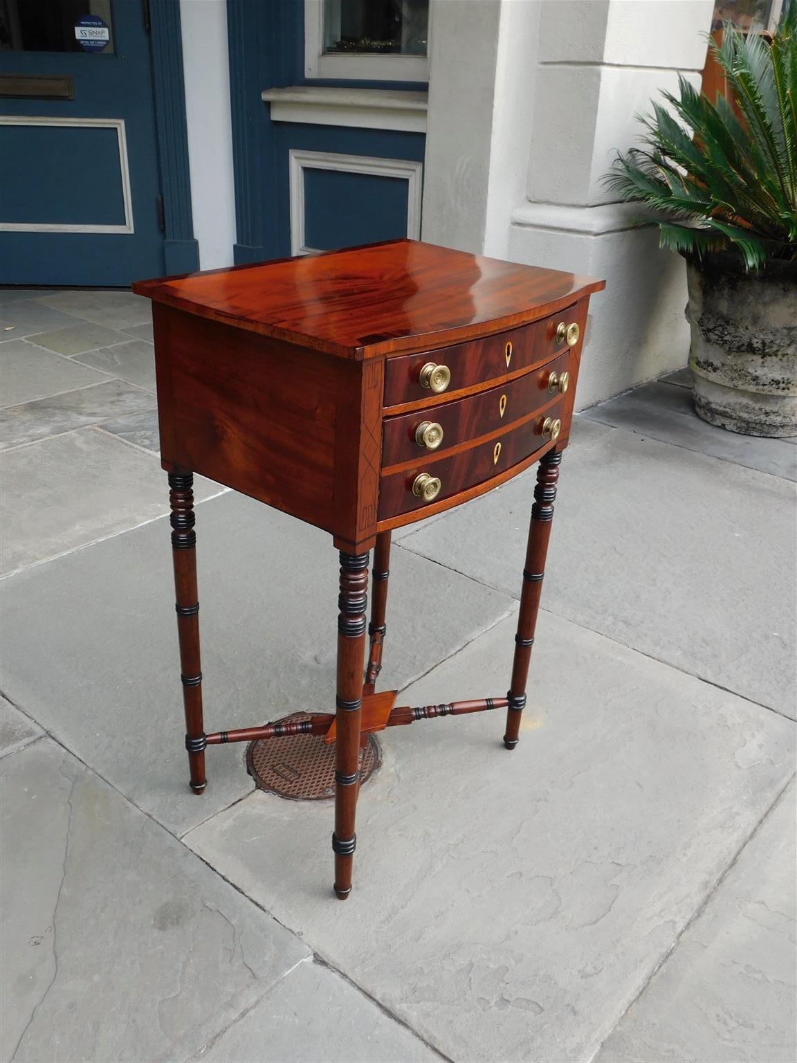 American Federal mahogany bow front two drawer work table with original circular foliate brasses, diamond bone escutcheons, ebony string inlays, kings wood cross banding, and resting on ebonized turned bulbous ringed legs with the original lower