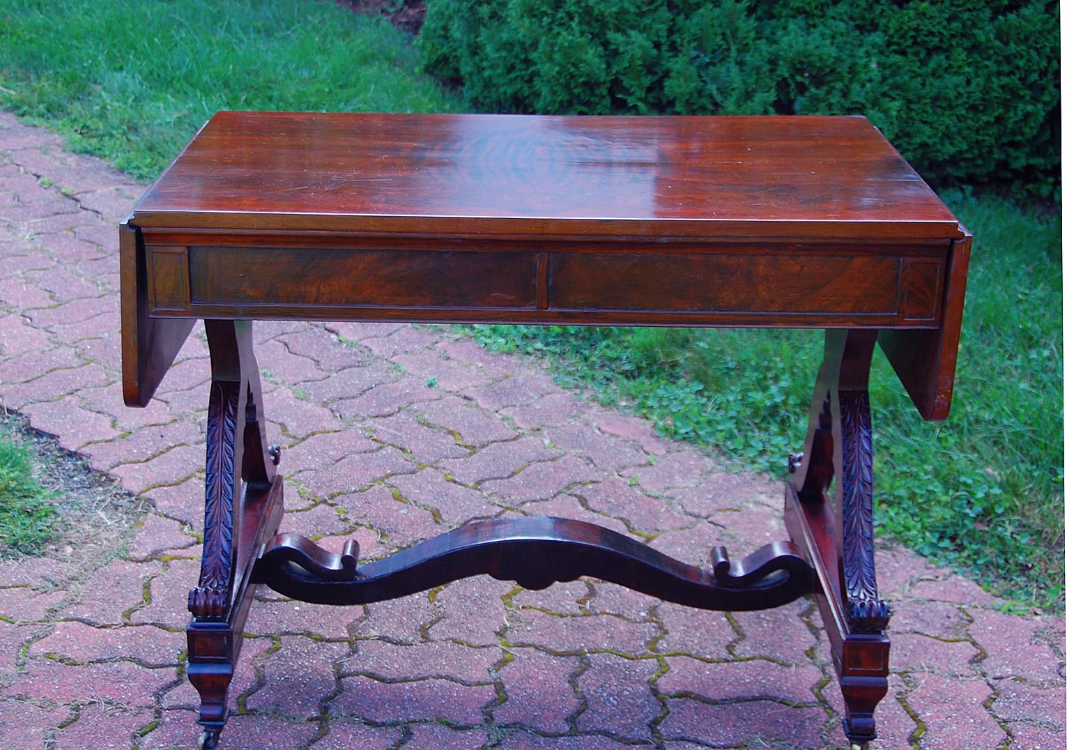 American early 19th century federal period rosewood sofa table with dropleaves, two drawers and unusual carved end supports. Acanthus leaves are carved down the curving end pedestals, a further unusual detail is the cross stretcher with undulating