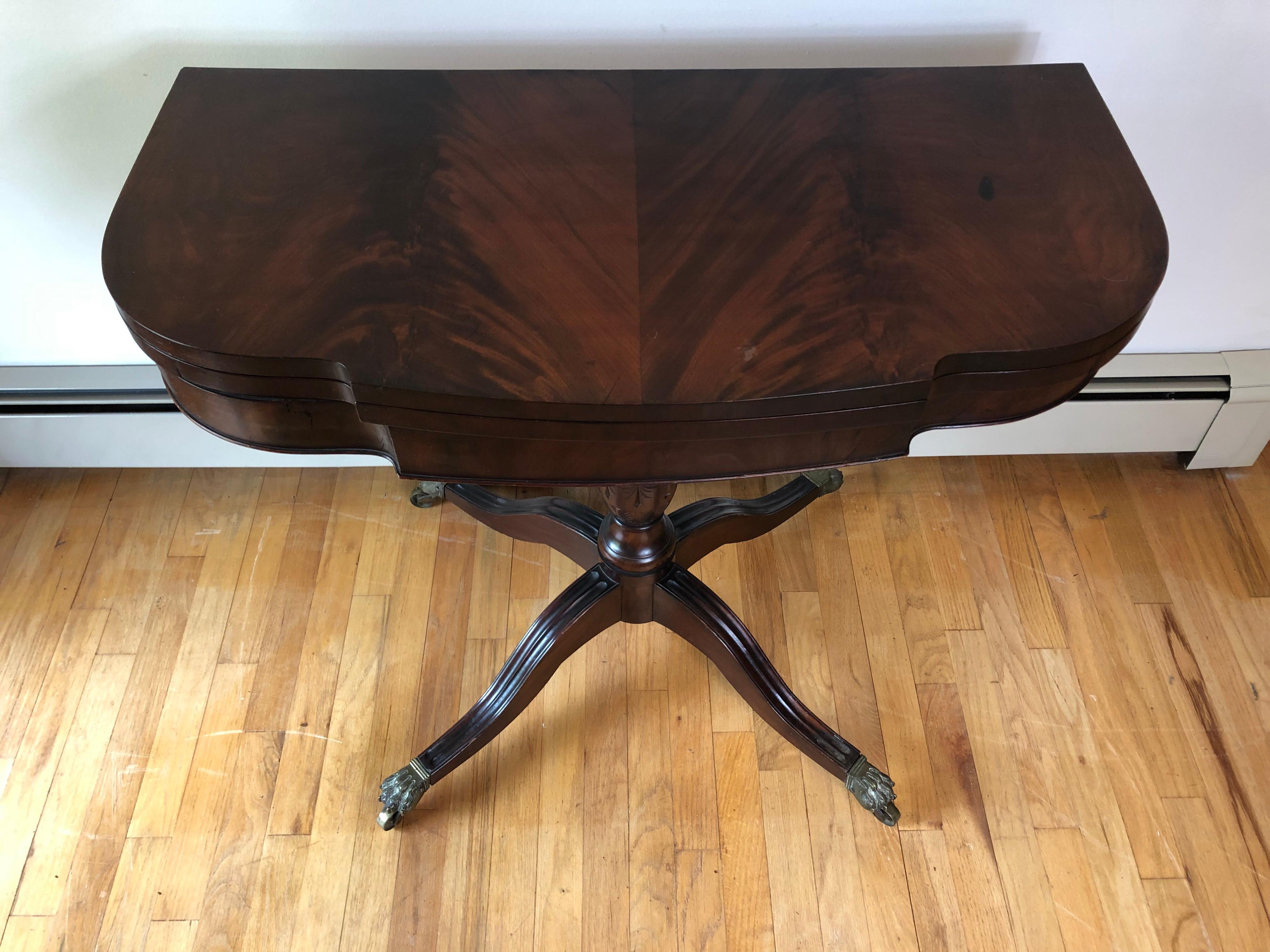 American Federal relief carved mahogany card table. The double-elliptical crotch mahogany book-matched veneered hinged top above a central pedestal with foliate and swag carved detail terminating on four fluted cabriole legs and brass lion paw cups