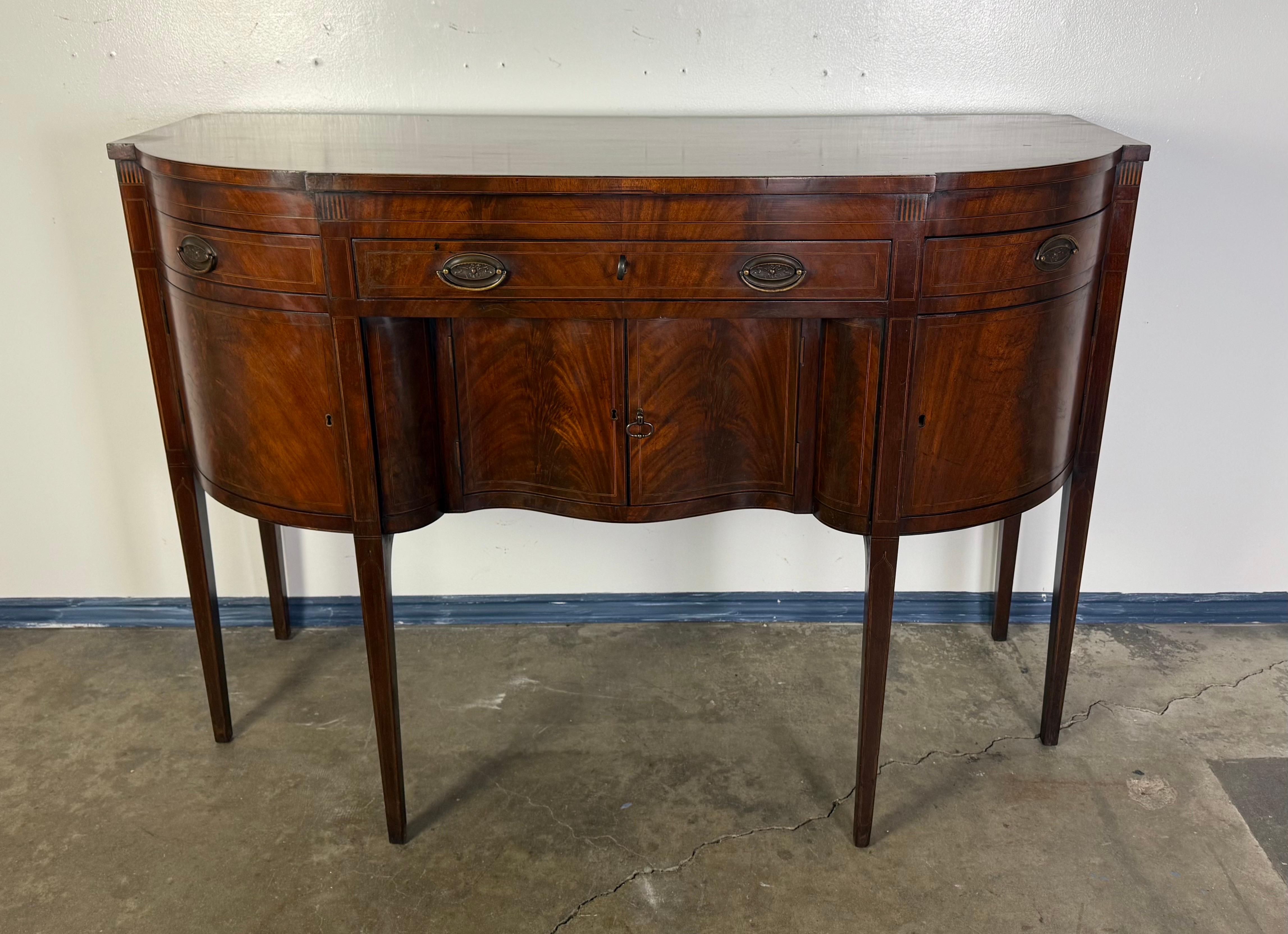 This is a finely crafted reproduction of a Federal-style sideboard or buffet, likely modeled after early American furniture designs from the late 18th to early 19th century. The piece features a serpentine front, tapered legs, and a rich mahogany