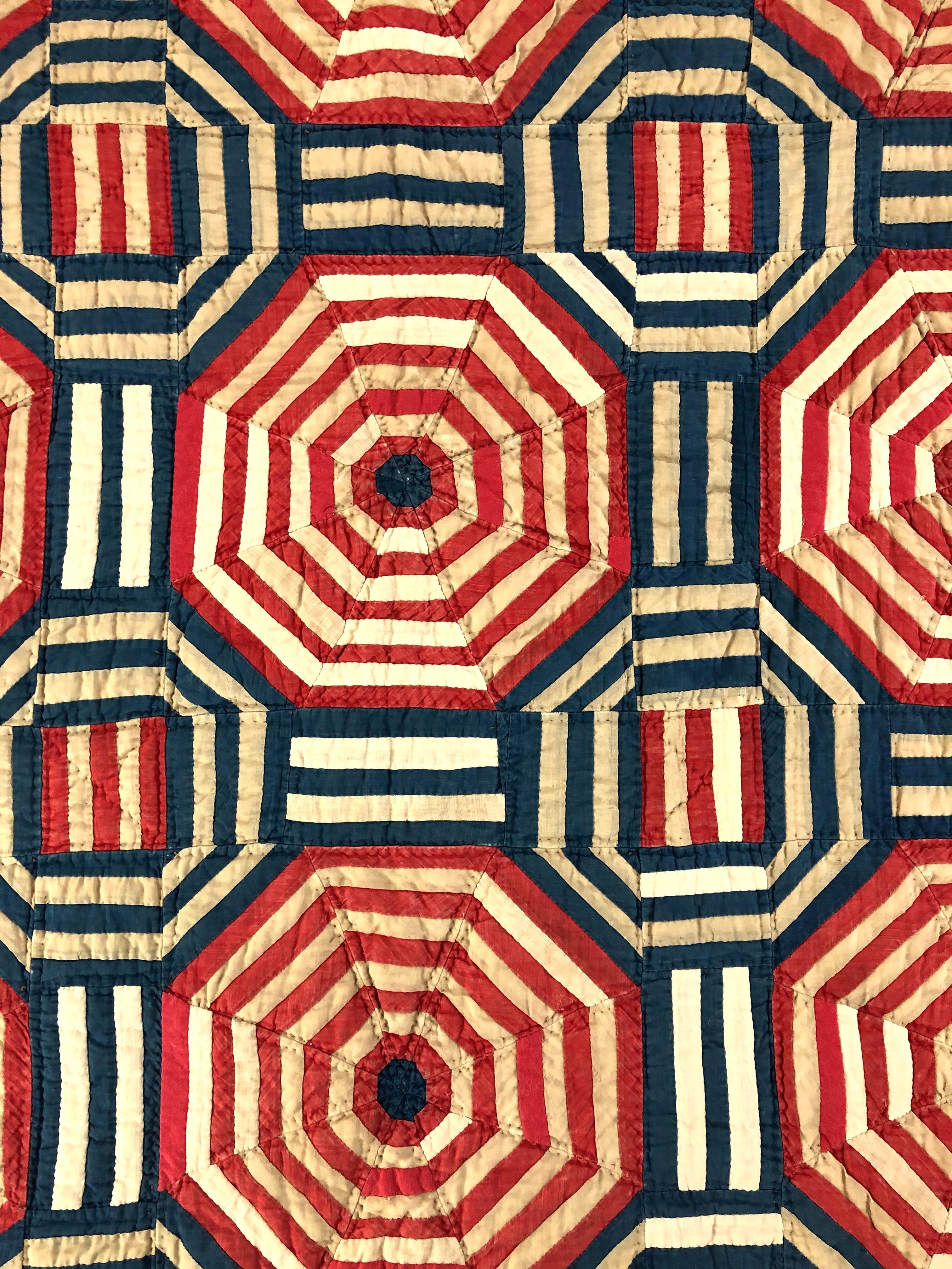 A strikingly graphic American Folk Art crib quilt wall hanging, in red, white and blue, composed of three rows of red and white striped hexagons interspersed with blue and white hexagons and red and white stripes, evocative of a modern Sol LeWitt or
