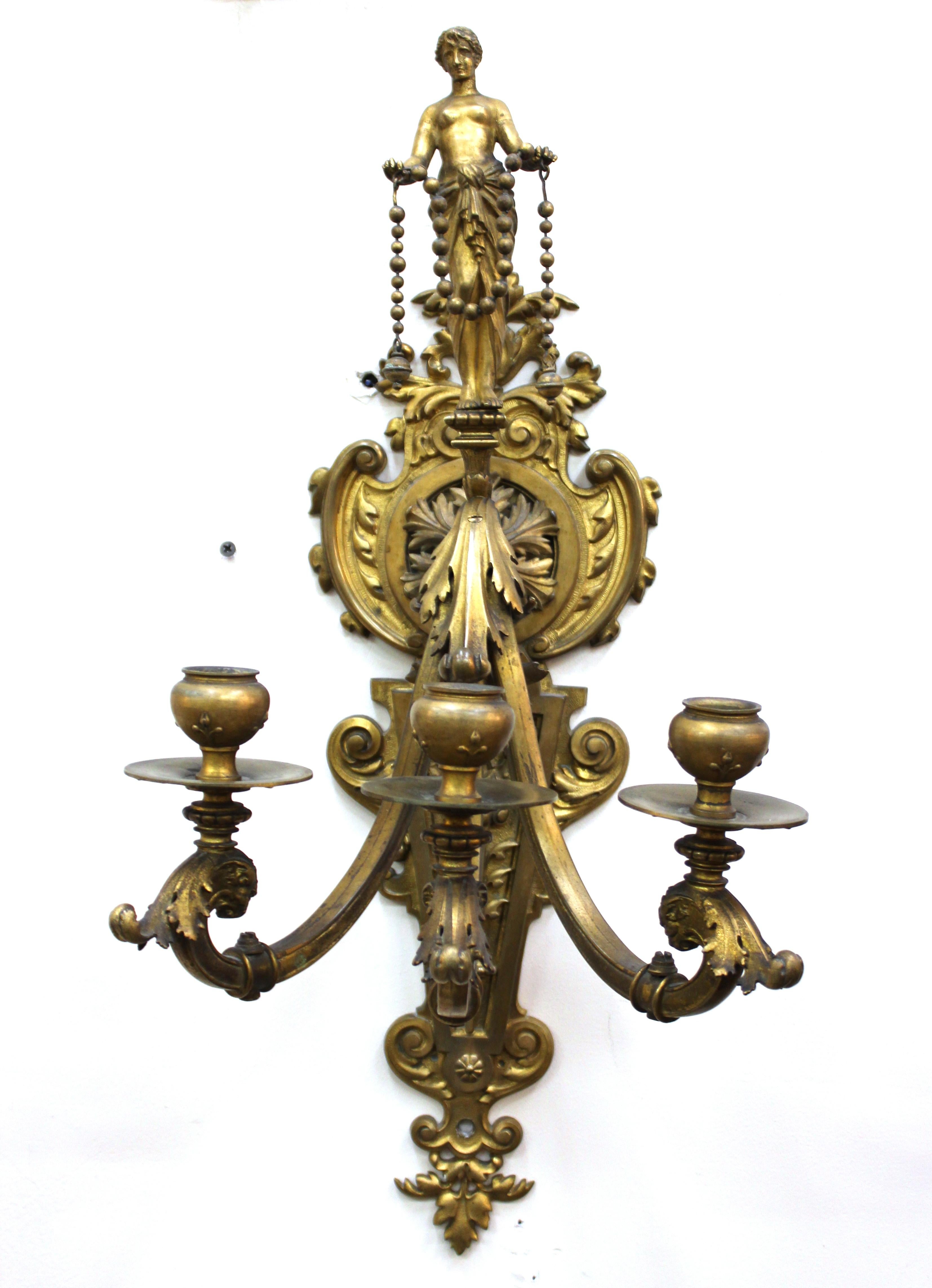 American Gilded Age period neoclassical style pair of wall candelabra sconces in gilt bronze with elaborate detailing and three candle holding arms each. Themed with foliage, grotesques and a nude standing allegory atop the scrolled arm. The pair