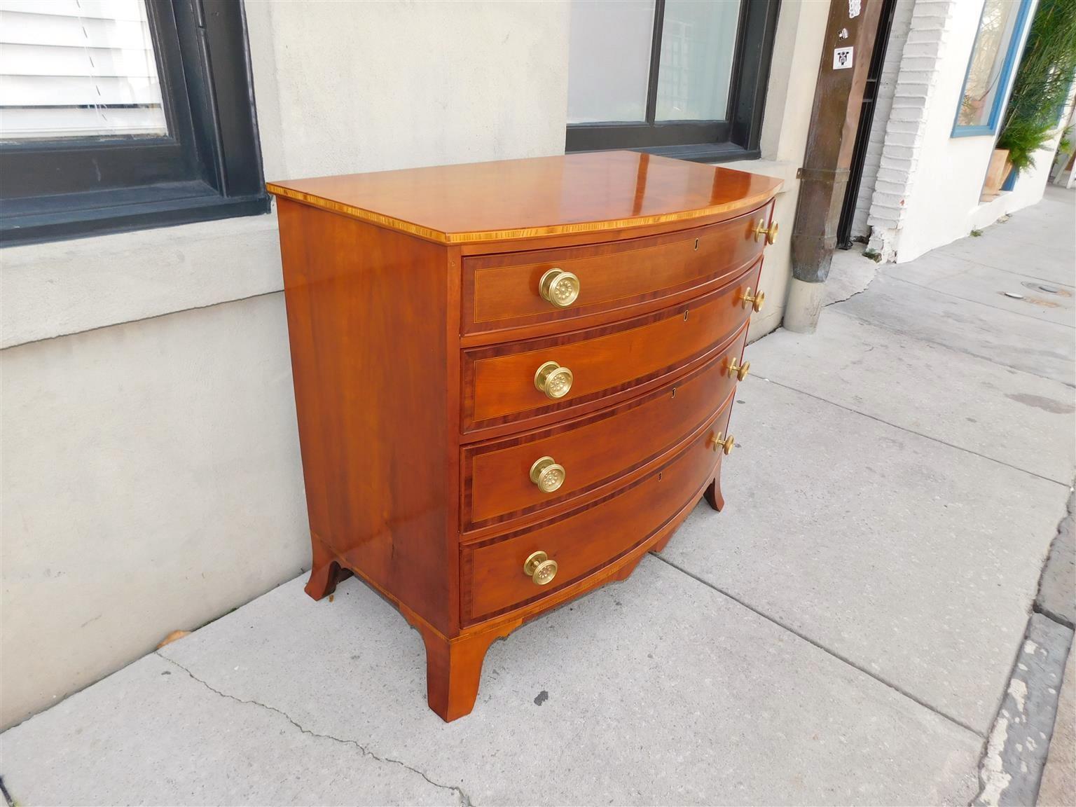 American Hepplewhite Cherry graduated bow front chest of drawers with satinwood border and string inlays, four cross banded mahogany drawer fronts, circular foliage brasses, central brass escutcheons with interior locks, carved scalloped skirts, and
