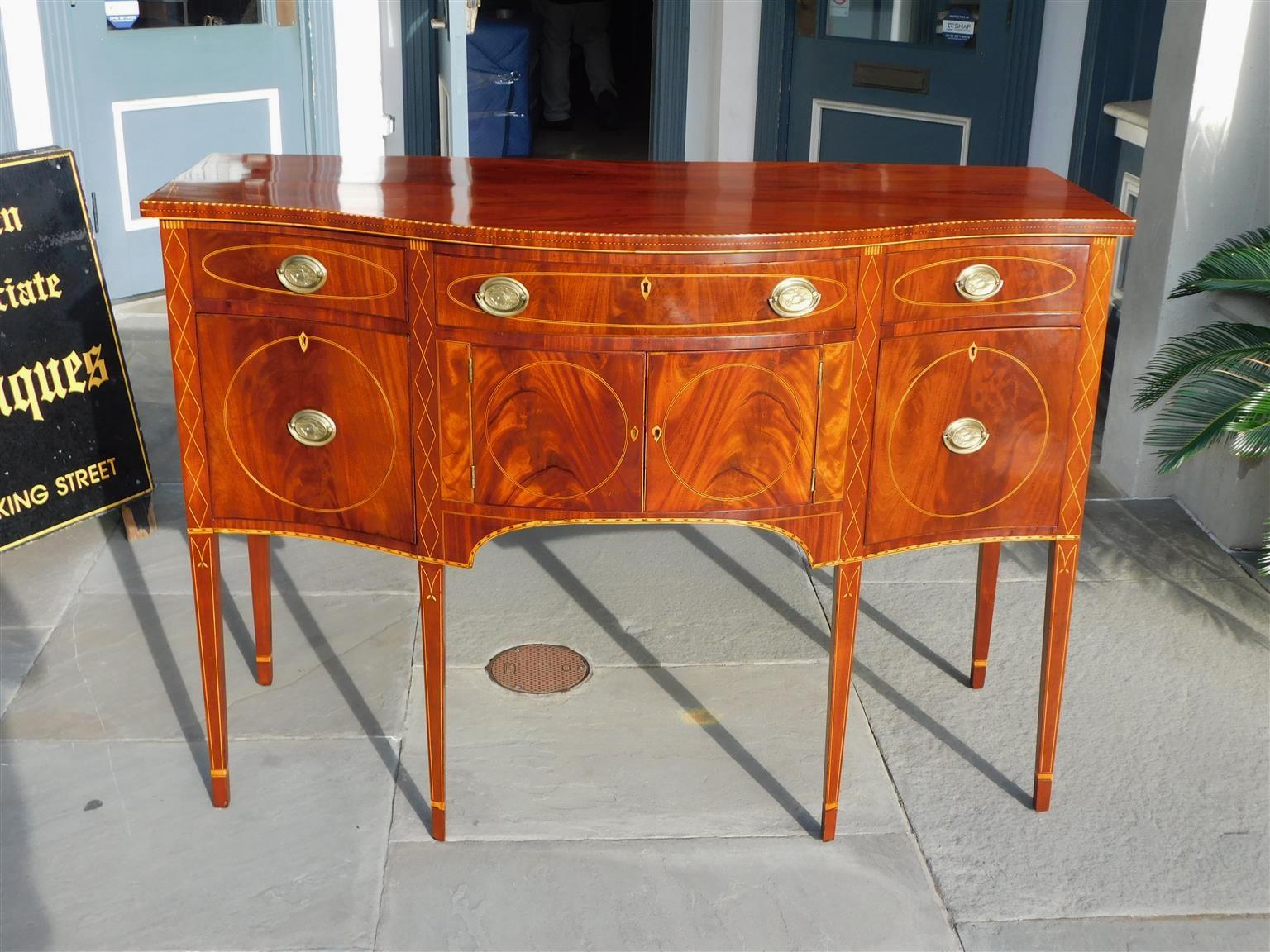 American Hepplewhite Mahogany serpentine sideboard with flanking oval satinwood string inlays, checkered inlays, period eagle oval brasses, and resting on the original satinwood string inlaid cuffed legs. William Whitehead, New York. Late 18th