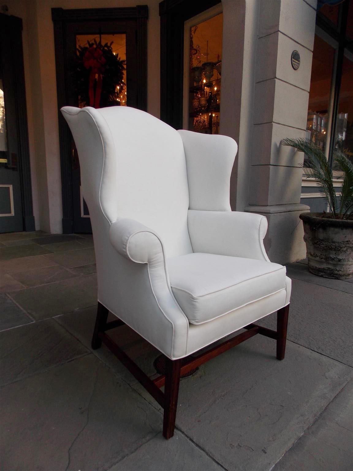 American Hepplewhite mahogany wing back chair with serpentine back, flared wings, flanking scrolled arms, removable seat cushion, and terminating on molded tapered squared legs with the original connecting stretchers. Secondary wood consist of White