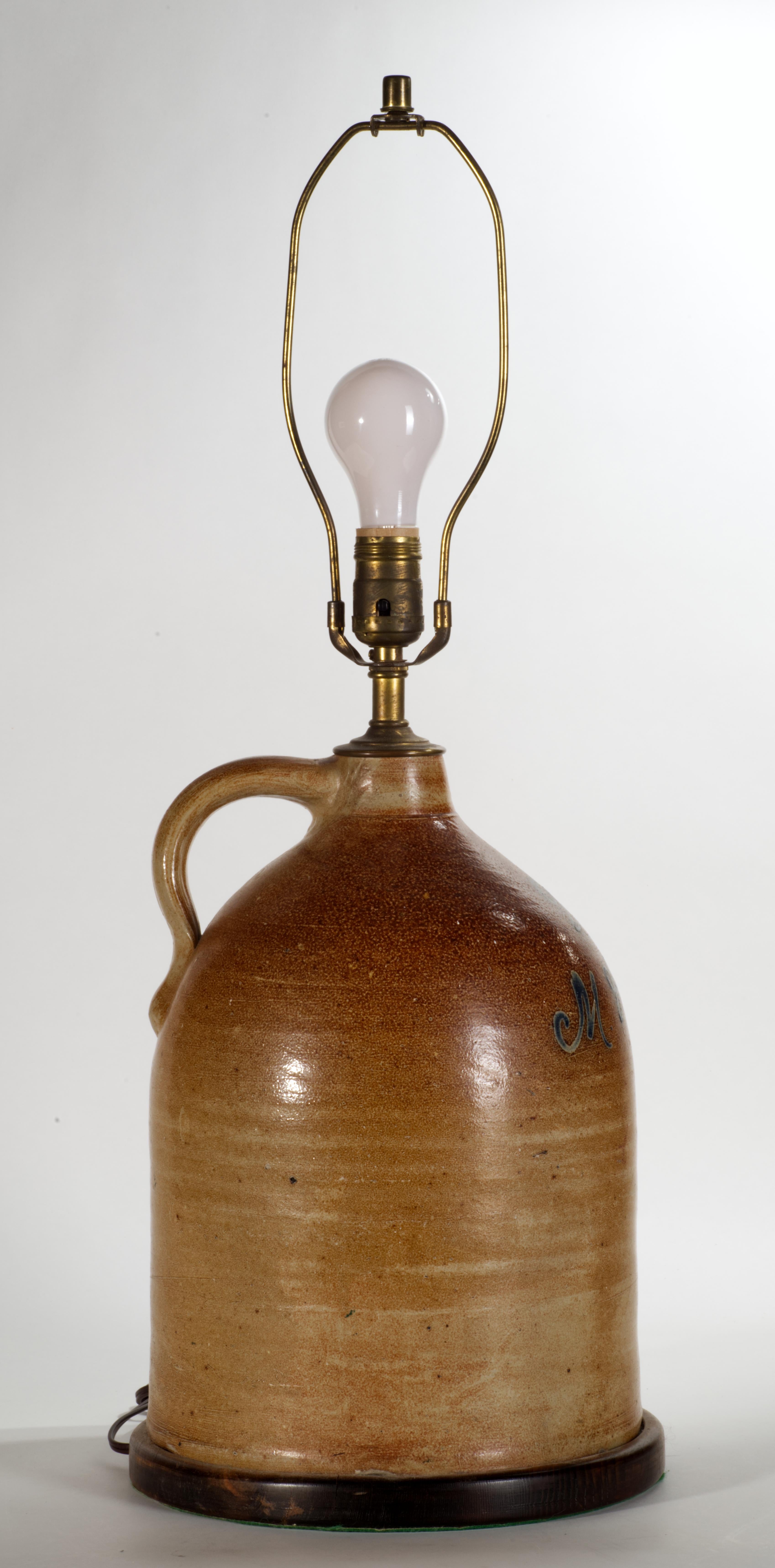 The antique brown glazed stoneware jug, converted into table lamp, features traditional shape with applied finger ring handle, rounded shoulder, and narrow neck; it is decorated with salt glaze in tone-on-tone ombre brown palette. The lamp has been